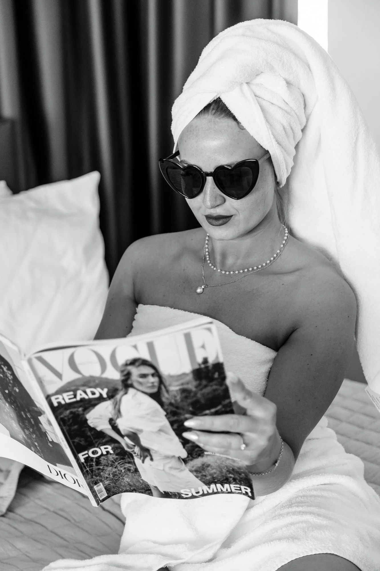 A woman with a towel wrapped around her head, wearing sunglasses, a pearl necklace, and a ring, sitting on a bed, reading a Vogue magazine.