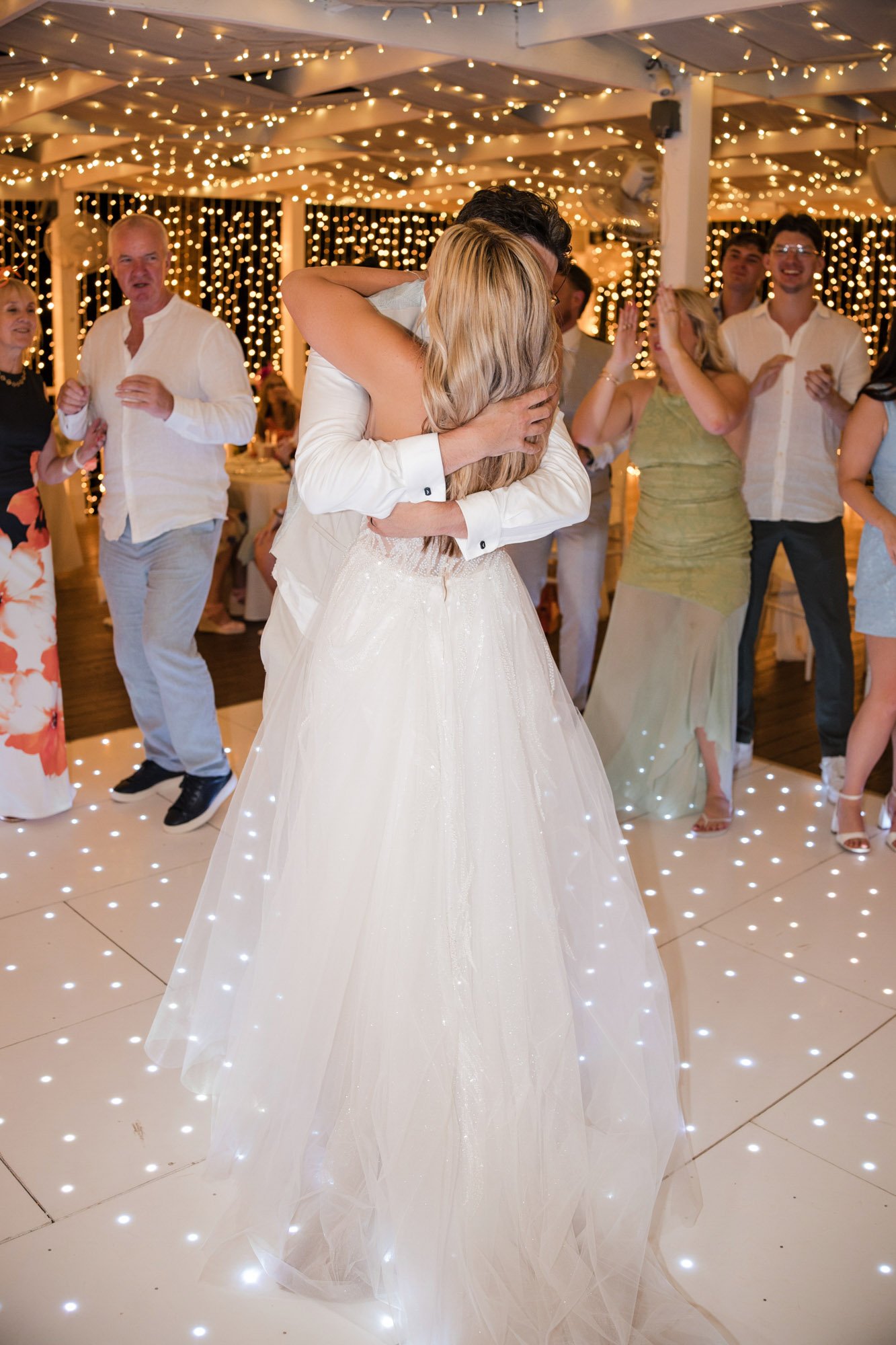 A bride and groom sharing a dance at their wedding reception with guests clapping and smiling around them, under fairy lights and a decorated ceiling.