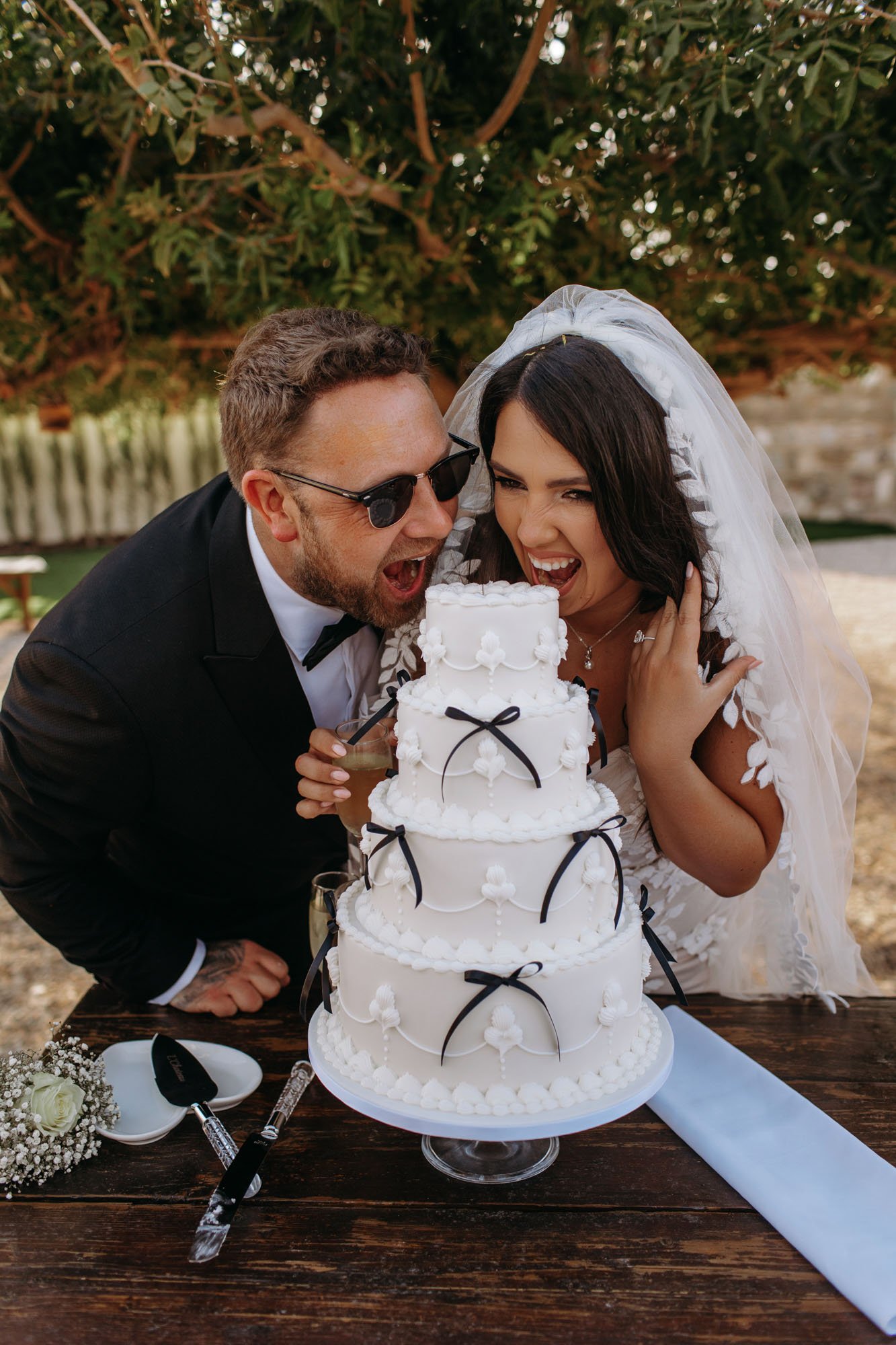 Bride and groom in wedding attire pretending to eat a tiered white wedding cake decorated with black ribbons and white flowers at an outdoor wedding celebration.