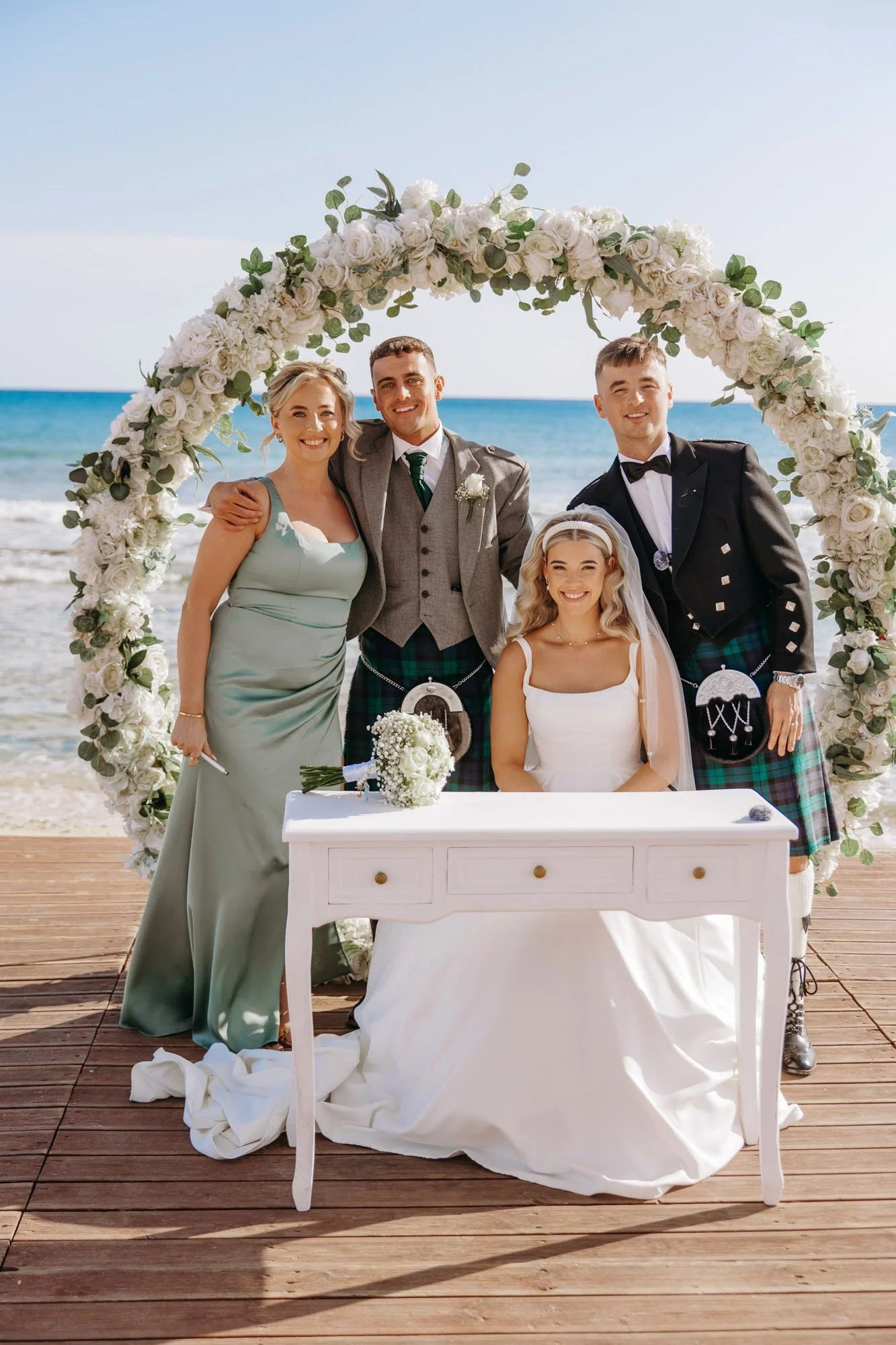 Group of five people at a beach wedding, standing behind a bride seated at a white table, with a floral arch and ocean in the background.