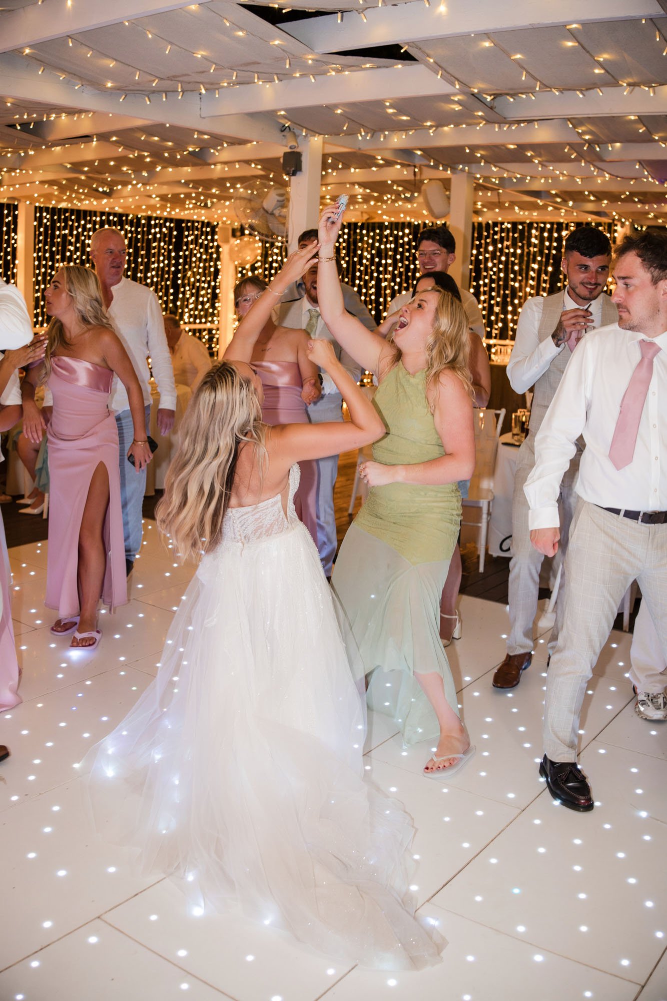 People dancing and celebrating at a wedding reception under string lights and fairy lights, with a bride in a white gown and guests in formal attire.