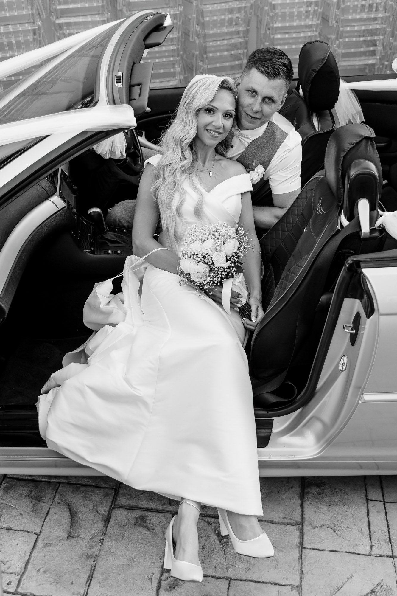 Black and white photo of a bride sitting in a convertible car with a bouquet of flowers, and a man standing beside her, both smiling, city skyline in the background.