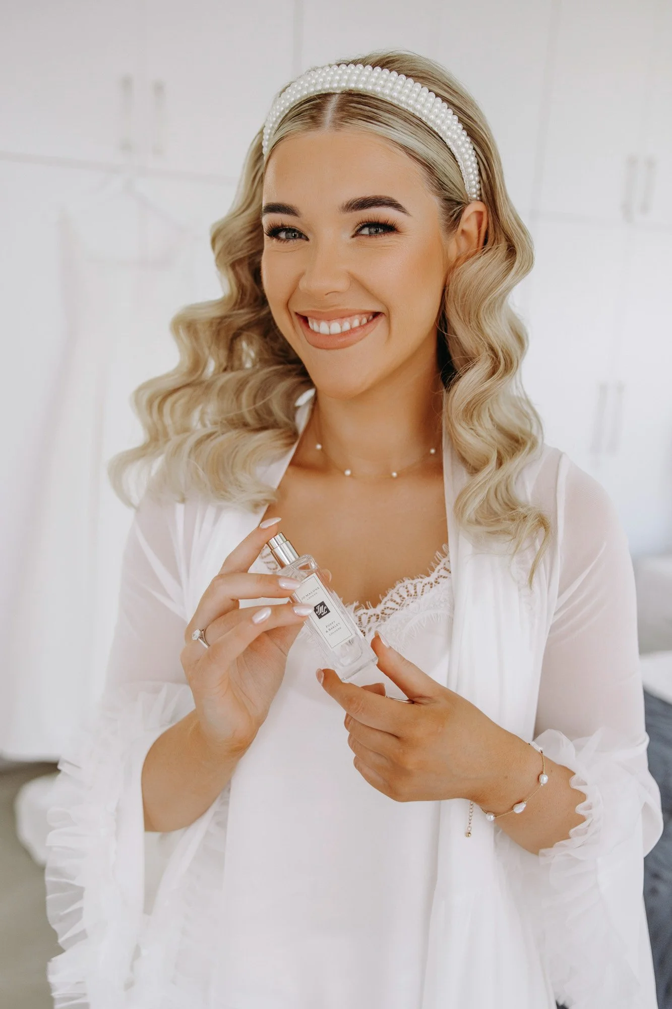 A woman in a white lace top and sheer white robe holds a small bottle of perfume, smiling and winking. She is wearing a pearl headband, pearl jewelry, and has styled, wavy blonde hair.