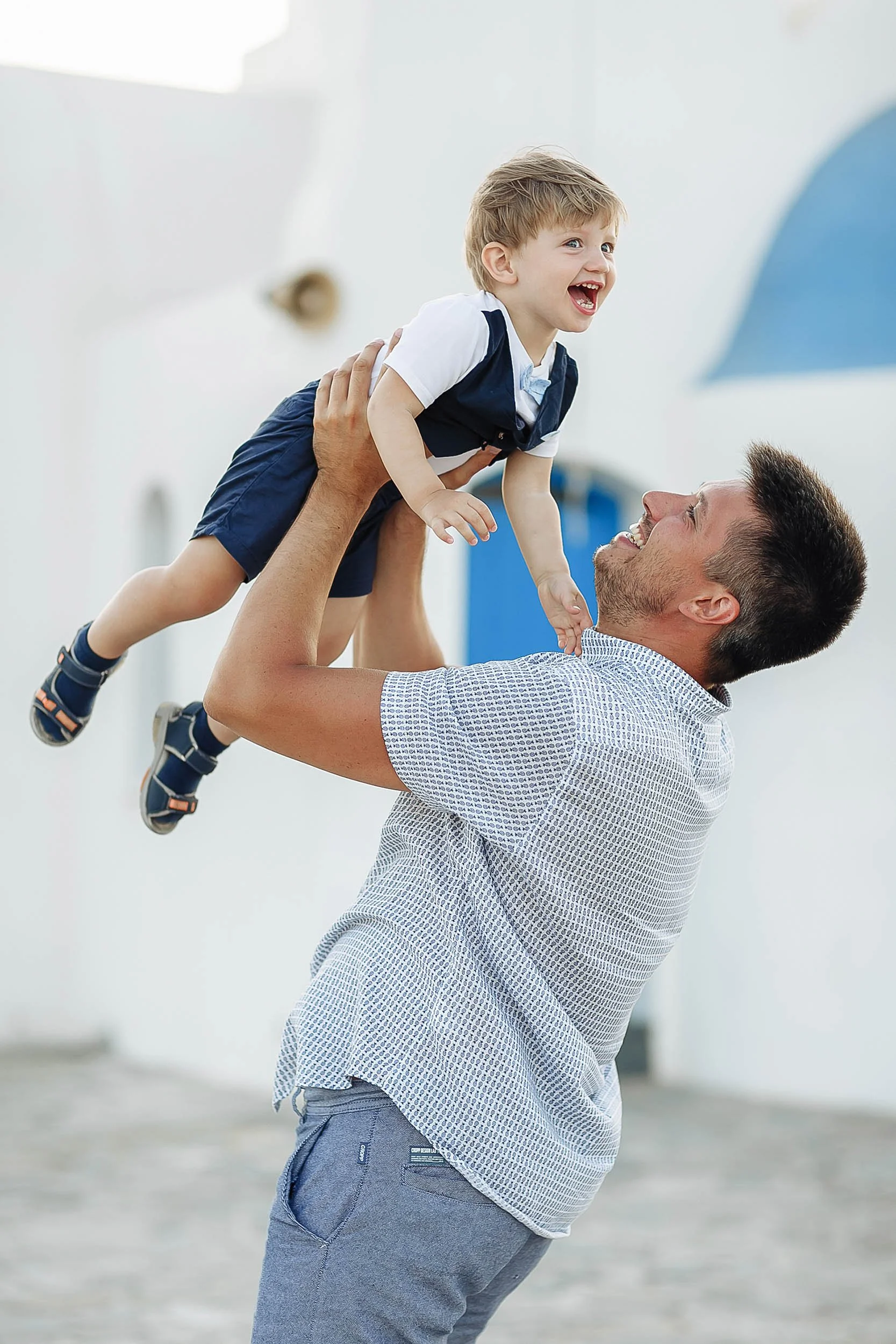 A man is lifting a laughing young boy with blond hair into the air outdoors on a bright day. The boy is wearing a white and blue shirt and shorts. The man is wearing a short-sleeved, patterned shirt and gray pants.
