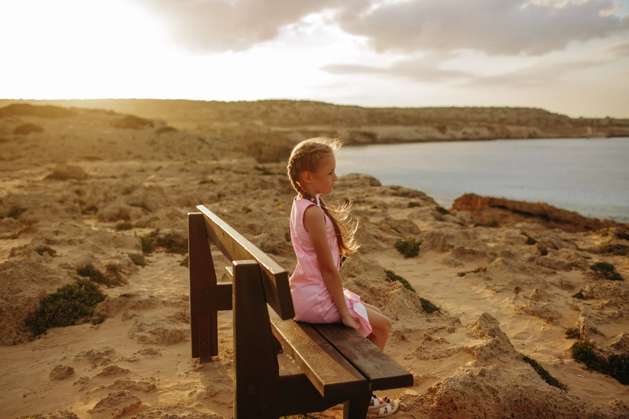 A young girl in a pink dress sitting on a wooden bench at the edge of a sandy beach, looking at the water, with a scenic view of a body of water, rocky shoreline, and cloudy sky during sunset.