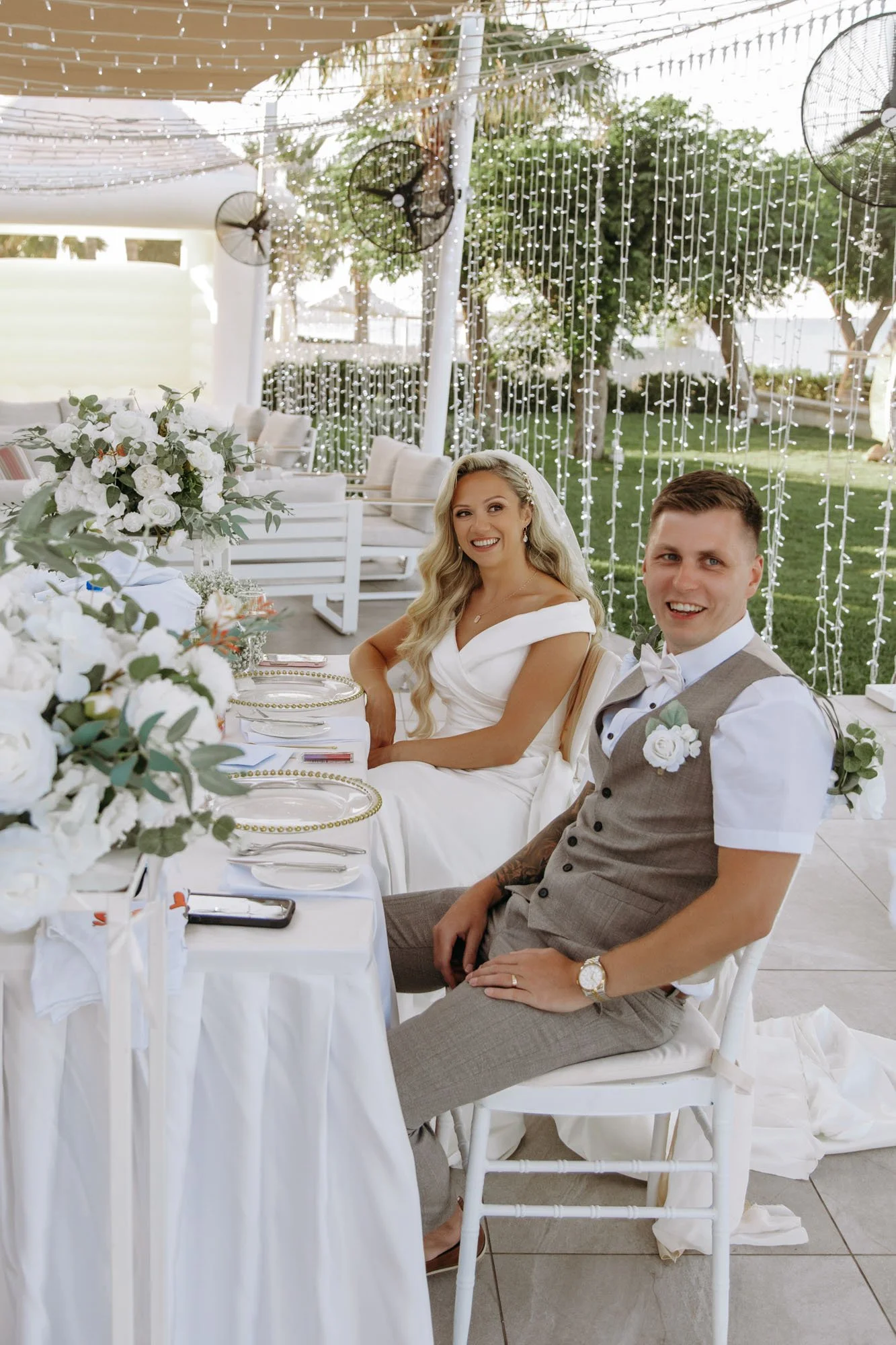 A bride and groom sitting at a wedding reception table outdoors, smiling, with decorated tables and string lights in the background.