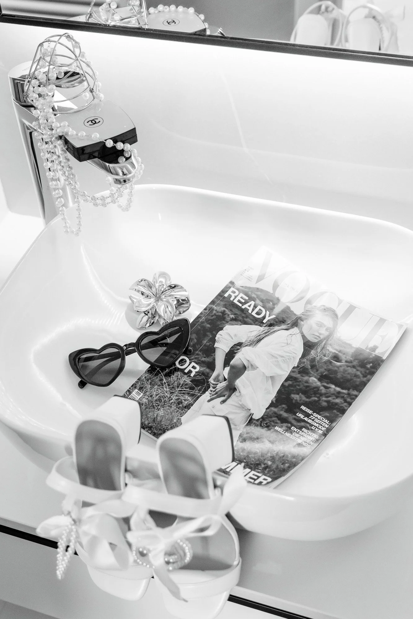 A bathroom sink with fashion accessories including sunglasses, pearl necklace, a flower-shaped hair clip, white high-heeled shoes with bows and pearl details, and a Vogue magazine with a model on the cover.