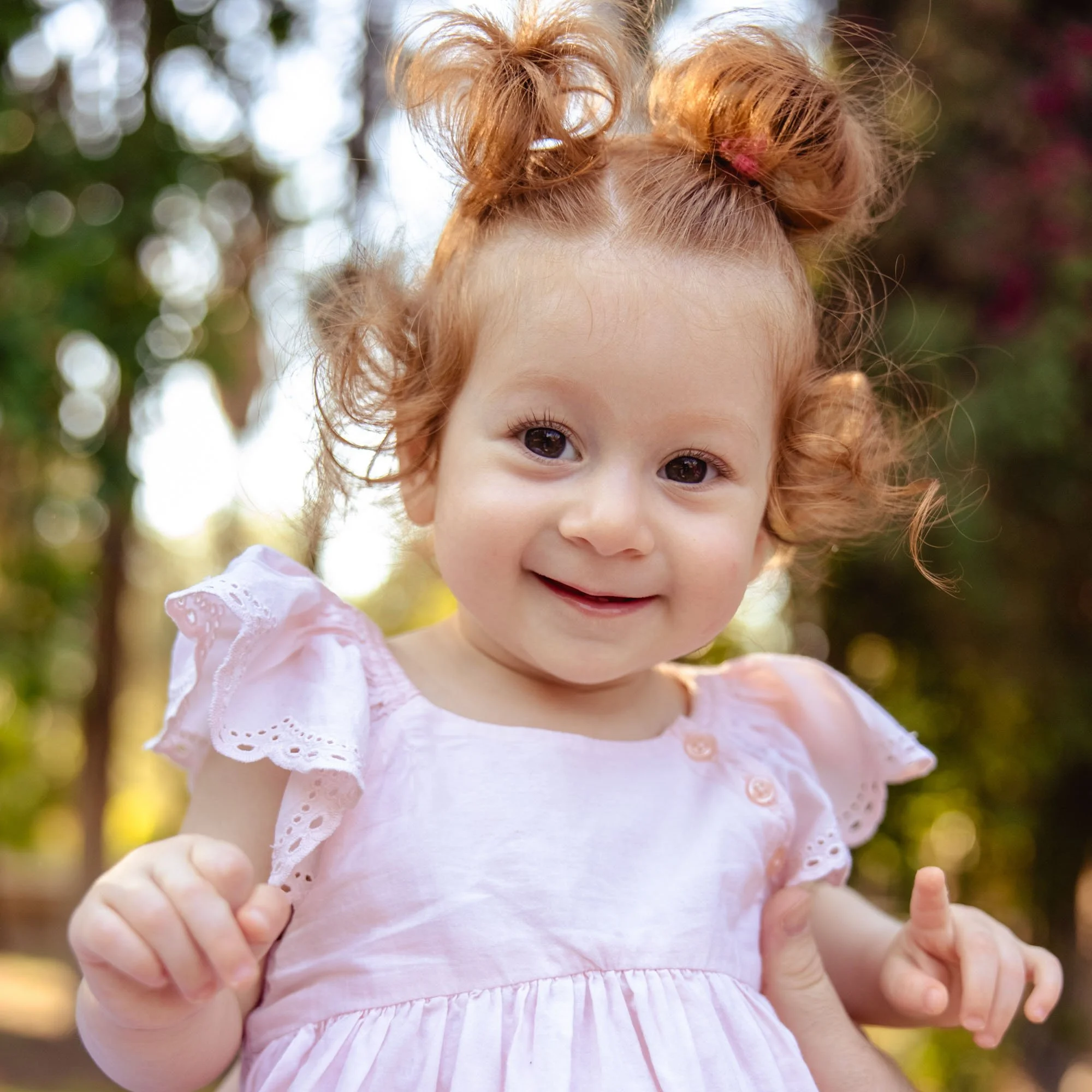 A young girl with curly red hair styled in two buns, smiling outdoors in a pink dress with ruffled sleeves, with a blurry background of greenery and sunlight.