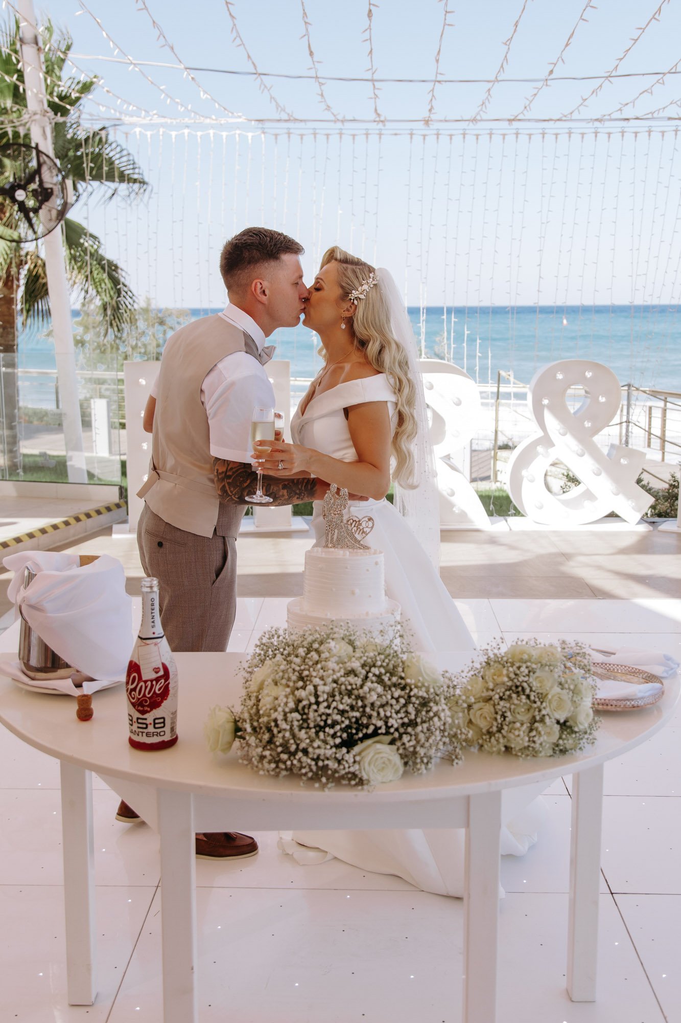A newlywed couple kisses during their wedding reception by the ocean. The bride and groom are holding glasses of champagne, standing behind a table with a wedding cake, flowers, and a bottle of drink. In the background, there are decorative lights an
