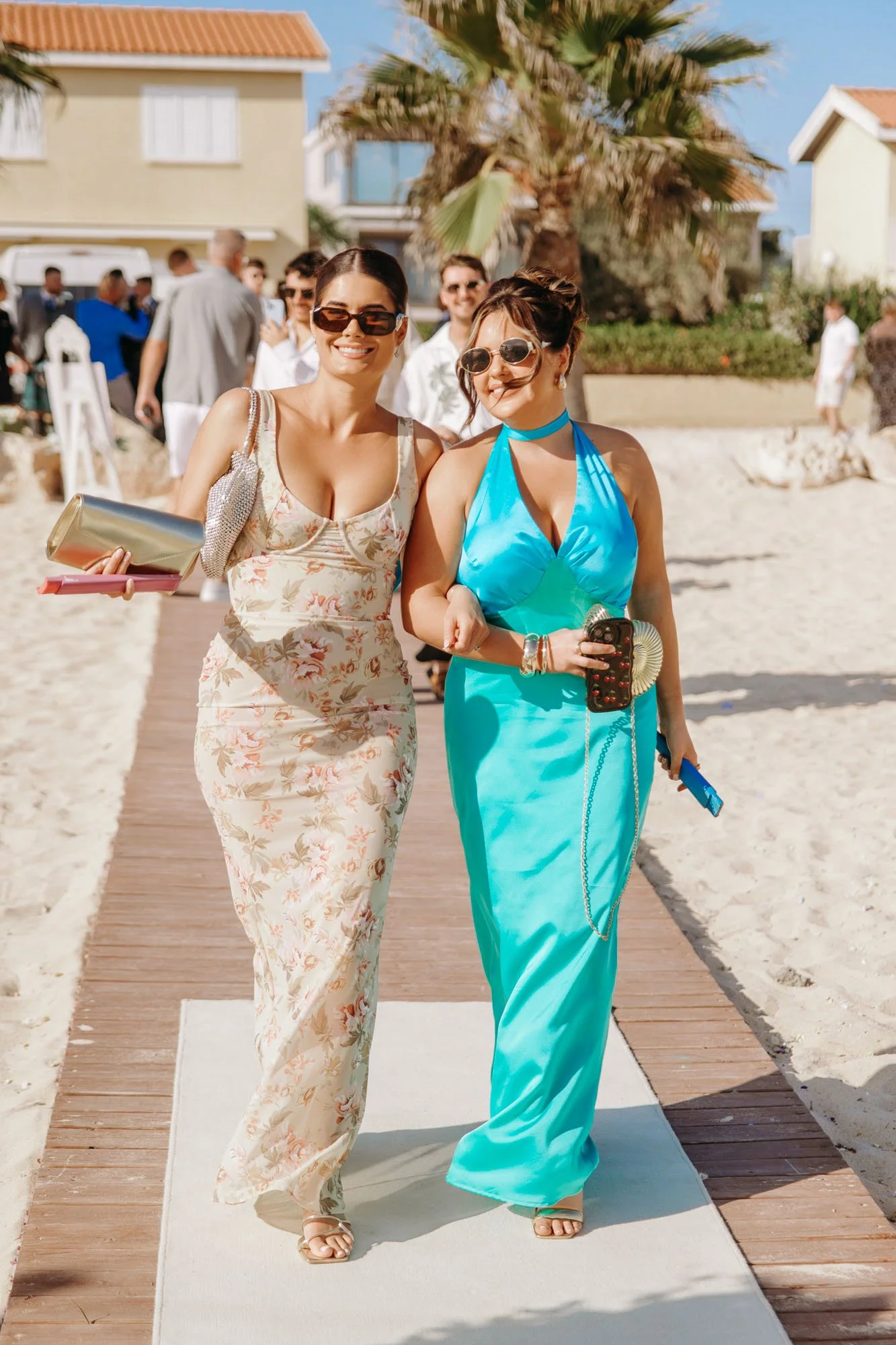 Two women walking arm-in-arm on a beach walkway, dressed in summery dresses and sunglasses, with a group of people and houses in the background, during a sunny day.