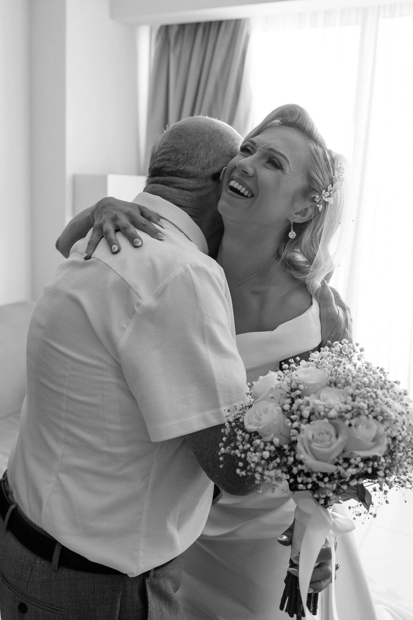 A black and white photo of a bride hugging an elderly man, likely her father, on her wedding day. The bride is smiling and holding a bouquet of roses and baby's breath.