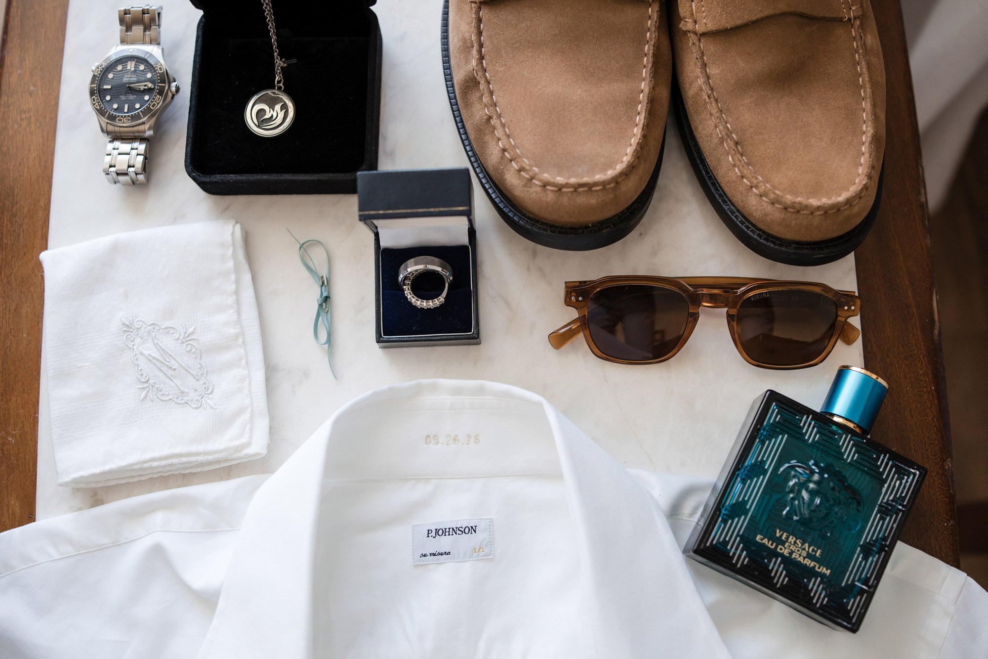 Collection of men's accessories and clothing arranged on a table, including a silver watch, a necklace with a pendant, a ring, a white handkerchief, a pair of tan sunglasses, a pair of tan shoes, a bottle of Versace Eau De Parfum, and clothing with a