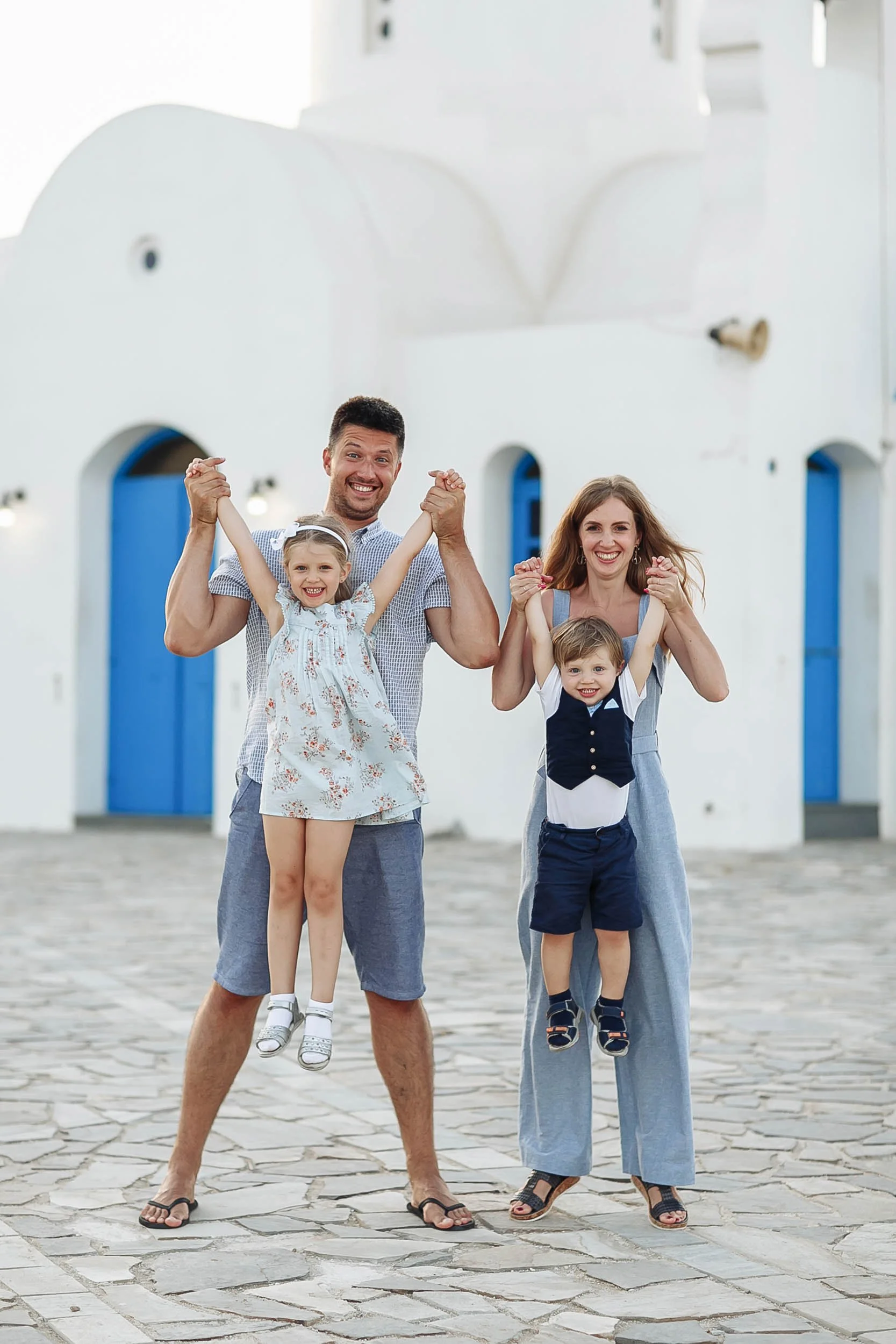 A smiling family of four standing outdoors in front of a white building with blue doors, with the father and young girl being lifted up by the mother and young boy, all holding hands and smiling.