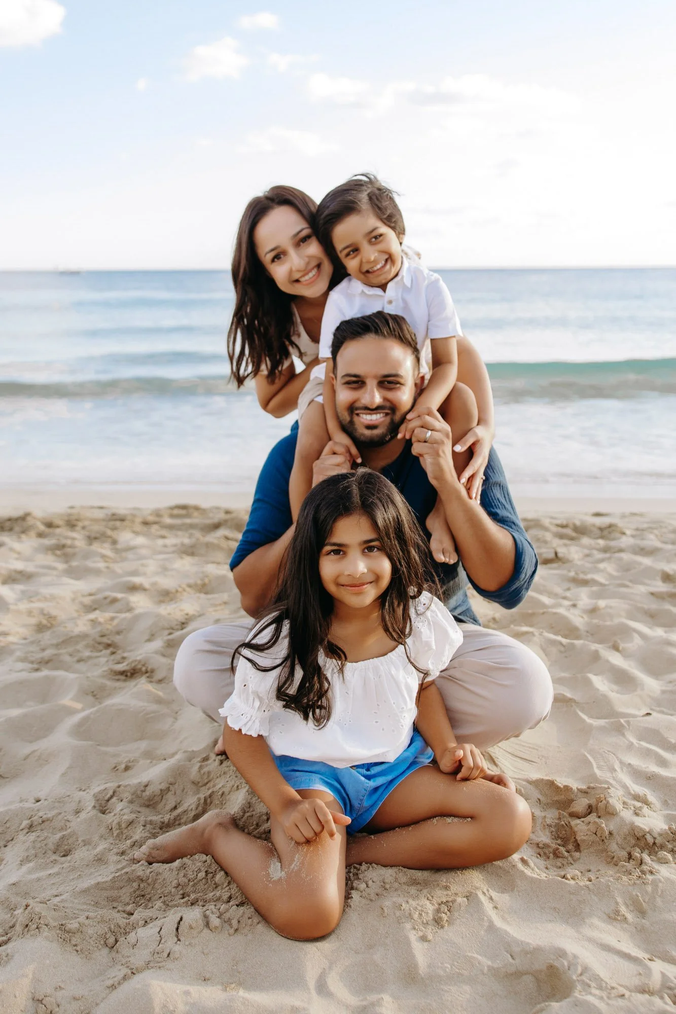 A family of five, including two children, sitting on the sandy beach with the ocean and sky in the background, smiling and posing for a photo.