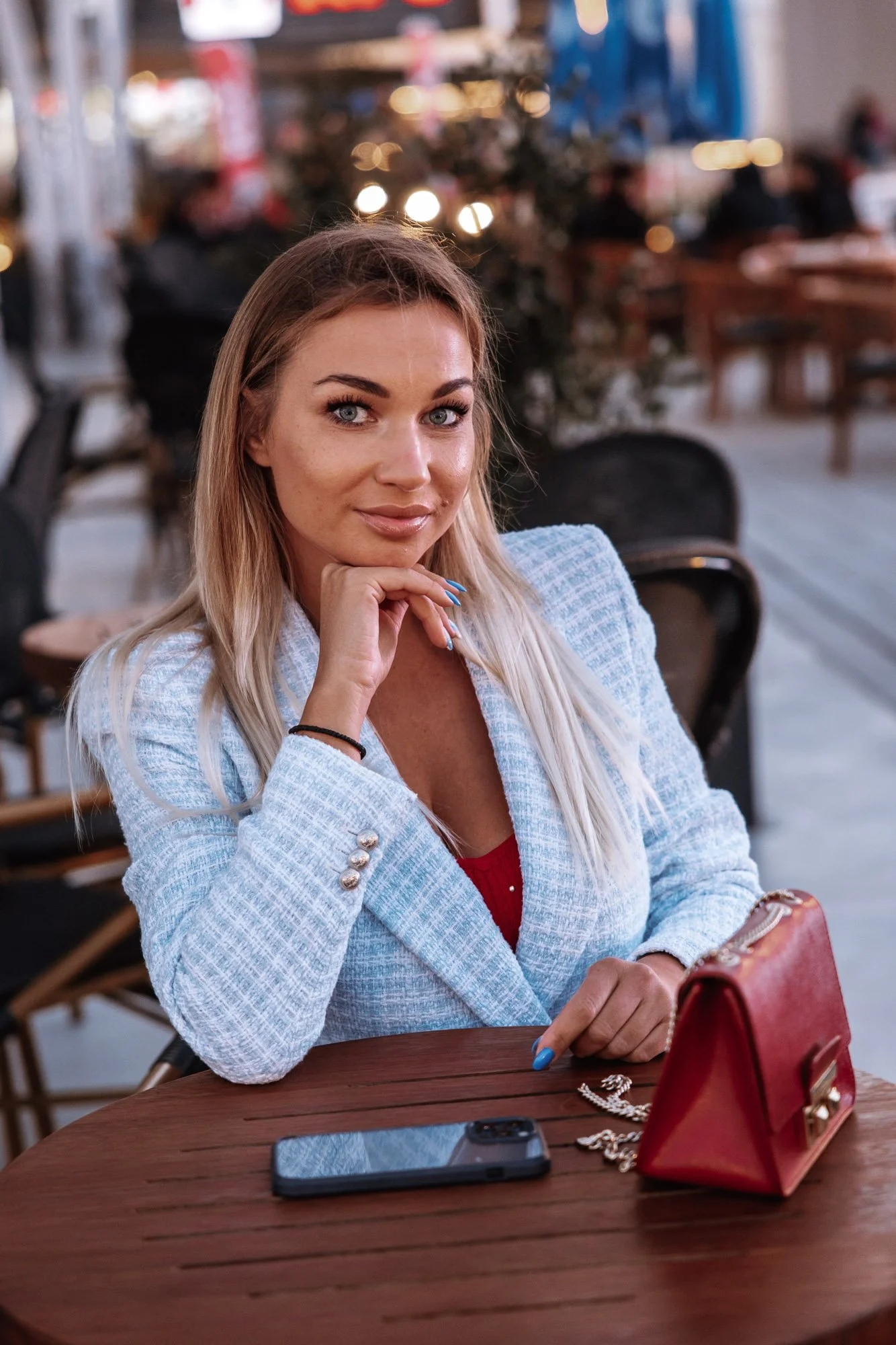 A woman with blonde hair and blue eyes sitting at a wooden table in a cozy indoor setting, wearing a light blue blazer and red top, with a red handbag, smartphone, and jewelry on the table, looking at the camera.