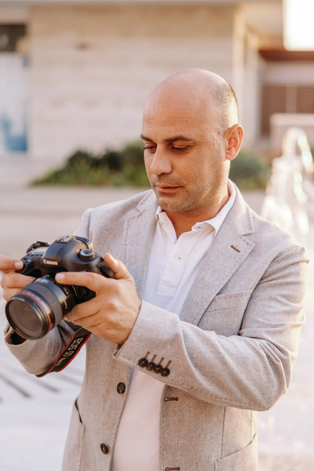 A man in a light gray blazer looking at a Canon camera in his hands outdoors.