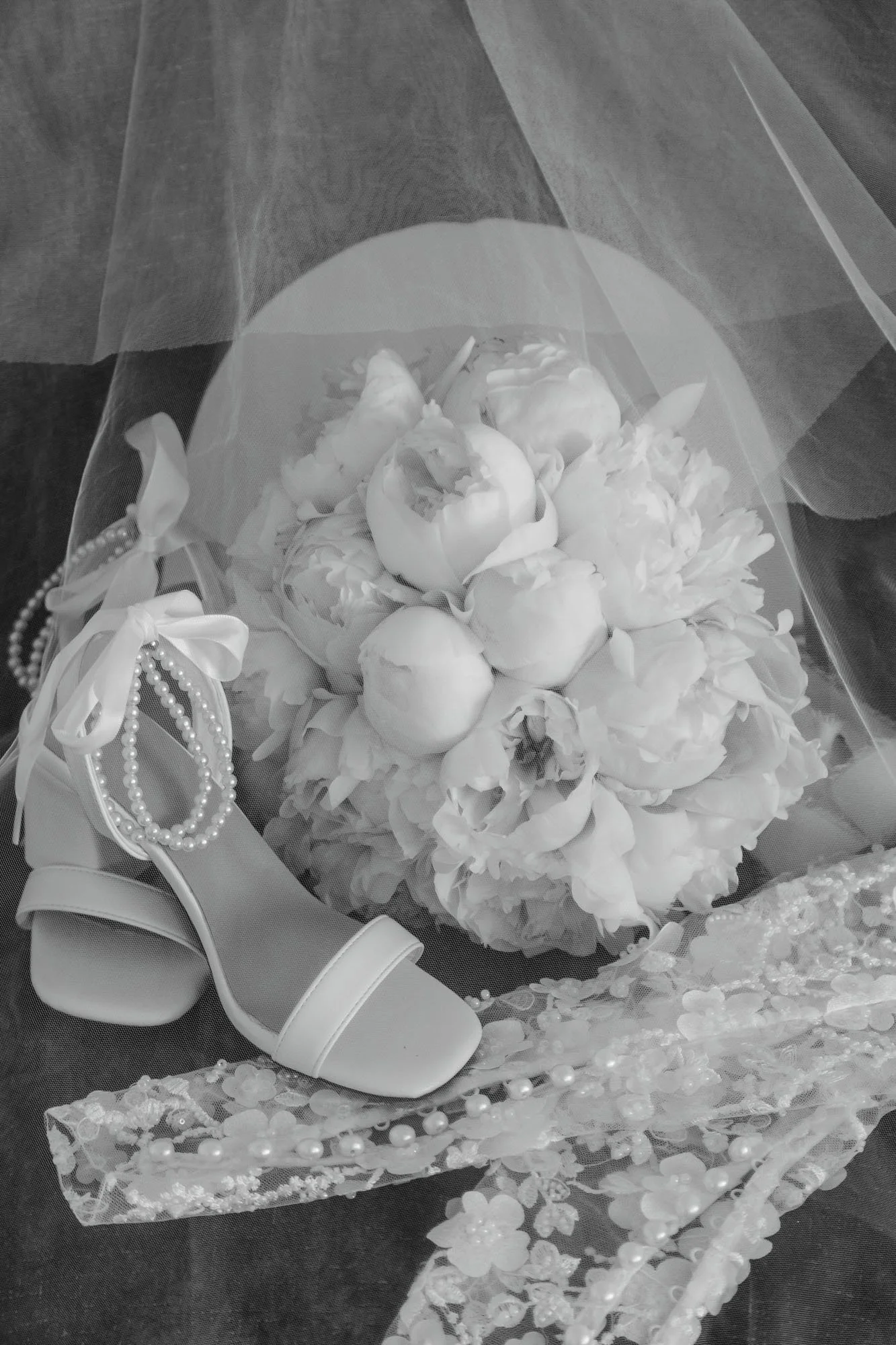 A bridal bouquet of peonies, a pair of white high-heeled shoes with ribbon and pearl decorations, and lace with pearls, all arranged on a dark surface, with a veil in the background.