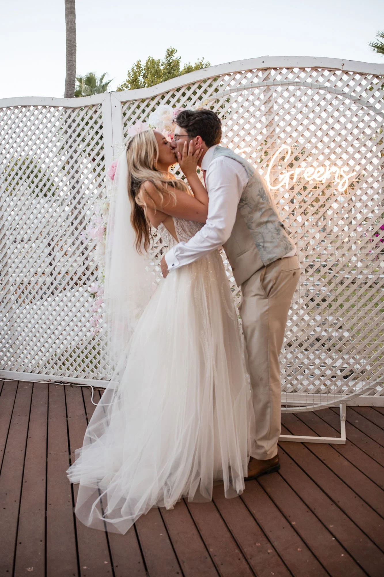 A bride and groom kissing at their wedding reception, with a white lattice backdrop and a neon "Greens" sign.