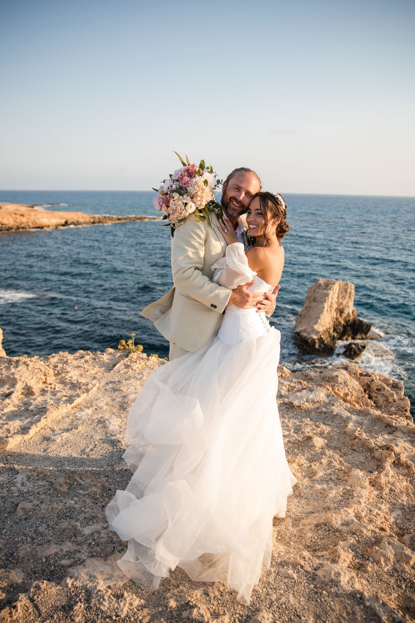 A newlywed couple embracing on a rocky coastline with the ocean and a clear sky in the background, dressed in wedding attire, with the bride holding a bouquet of flowers.