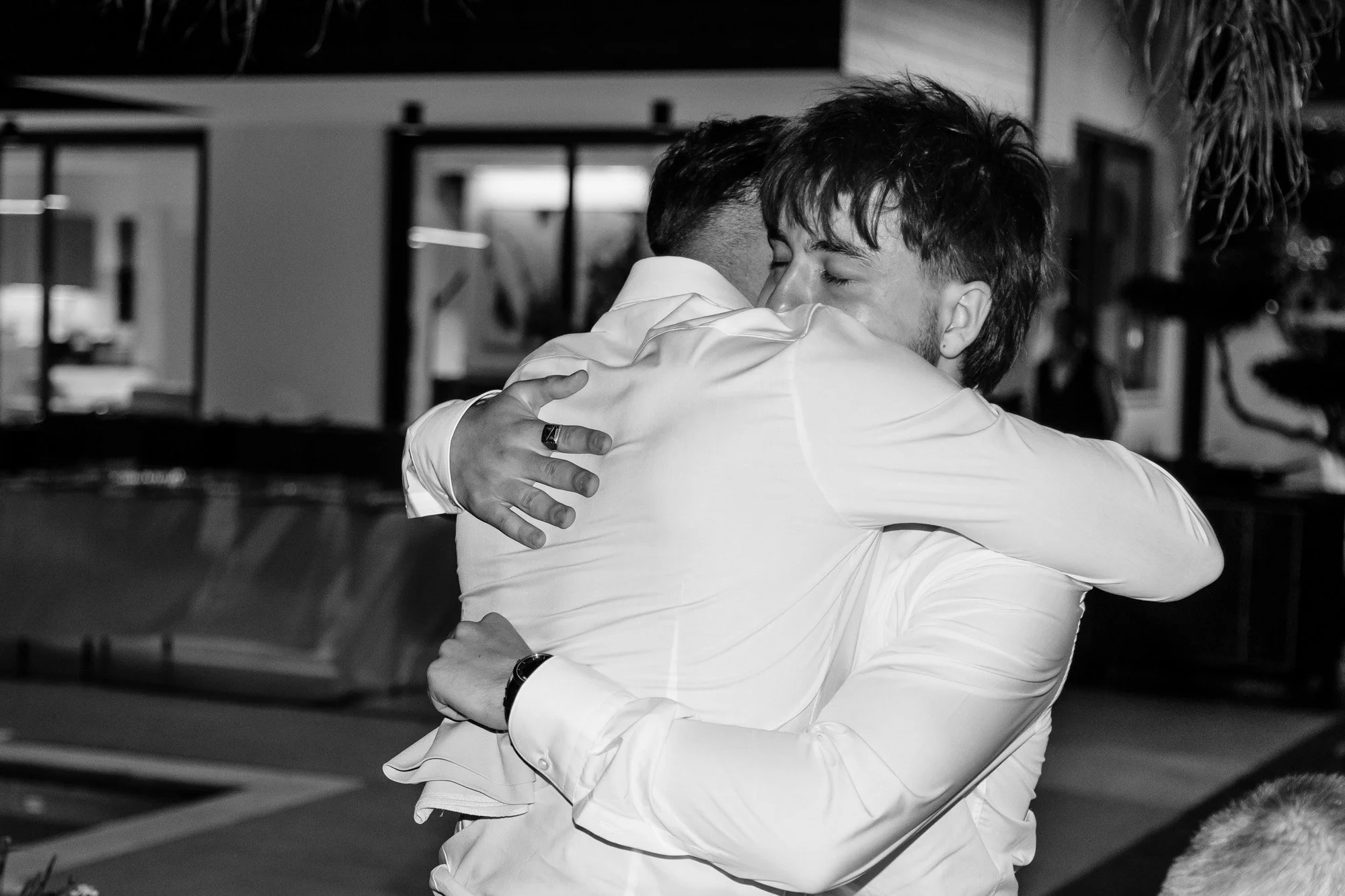 Two men hugging warmly, smiling with eyes closed. One of them is wearing a white dress shirt, and the other is dressed in a dark suit with a ring on his finger.