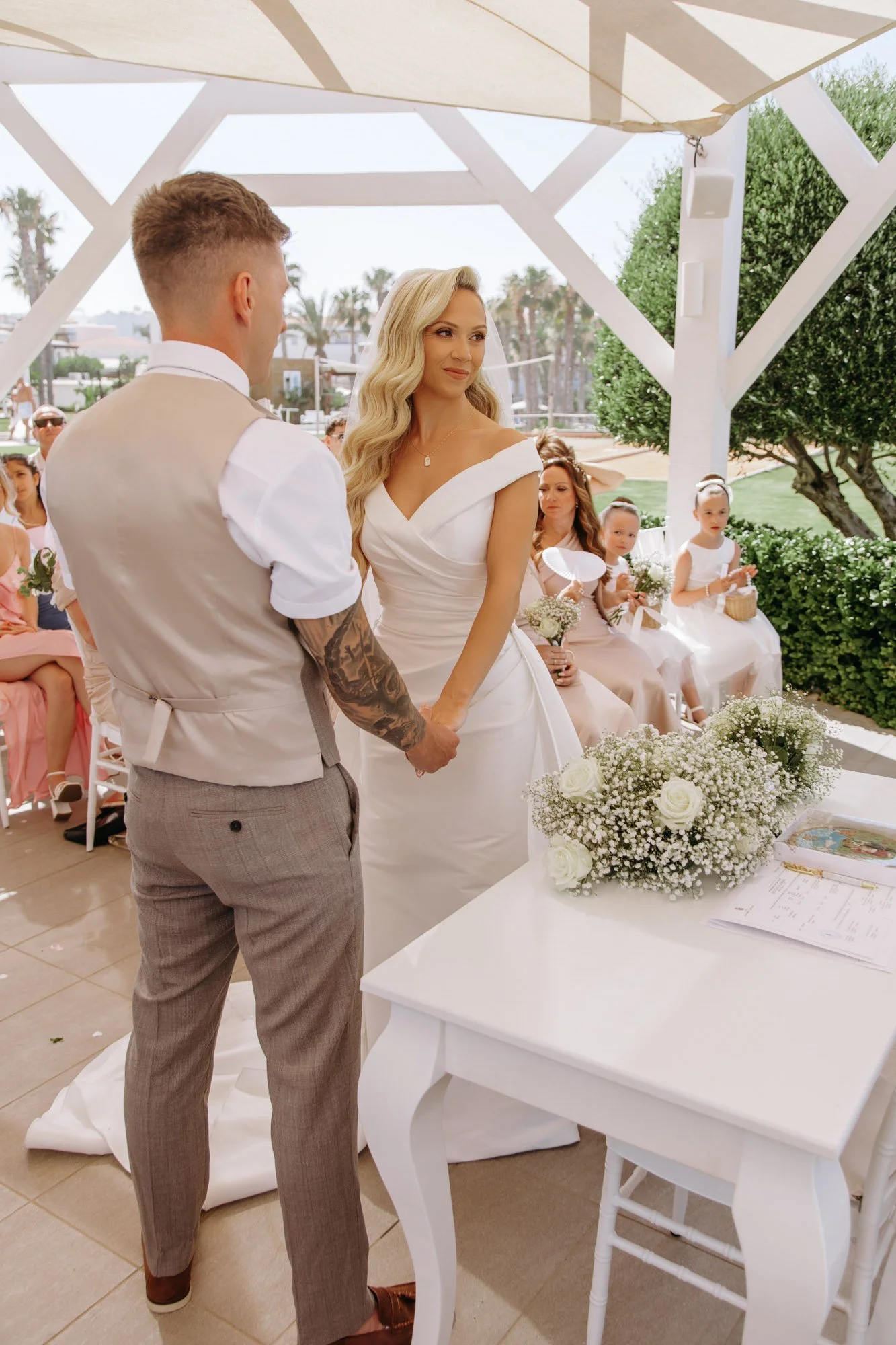 A wedding ceremony outdoors under a white canopy, with a bride and groom holding hands, surrounded by seated bridesmaids and guests.
