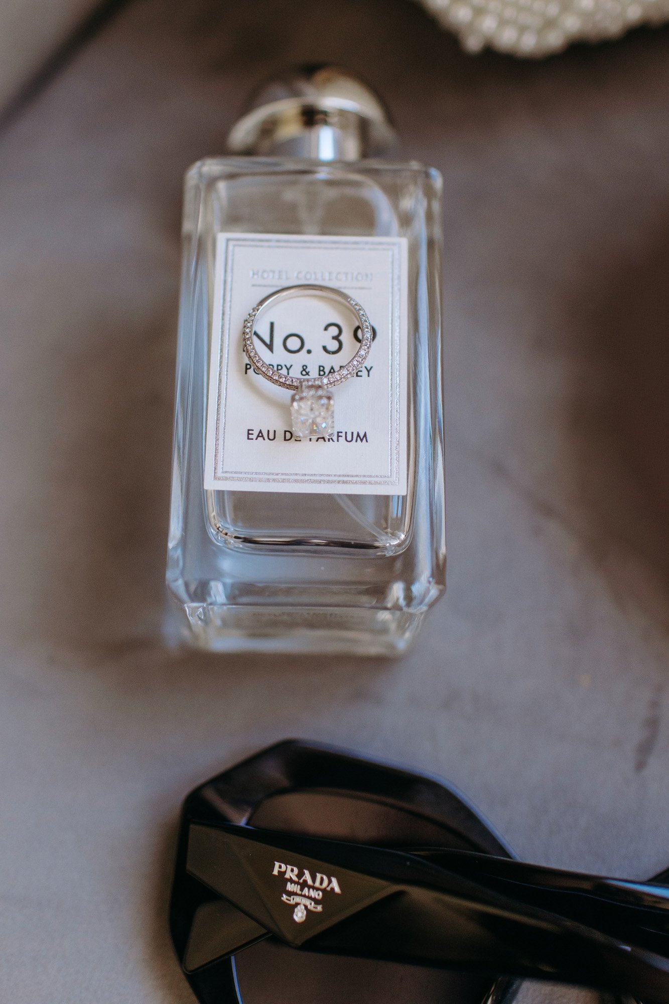 A bottle of hotel collection perfume with a ring on top of it, and a pair of black Prada sunglasses nearby.