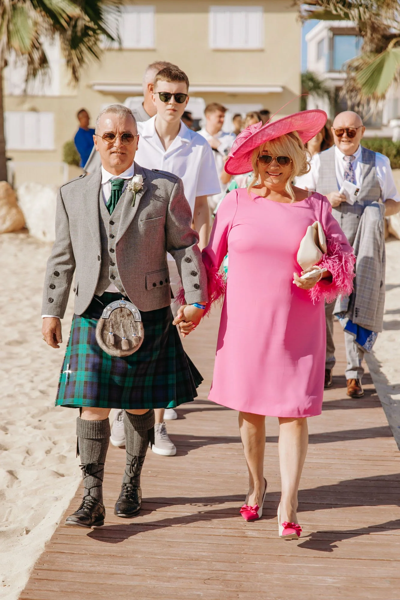 A group of people walking on a wooden path on a beach, with two in the front holding hands, dressed in formal and colorful attire, suggesting they are attending a wedding or special event.