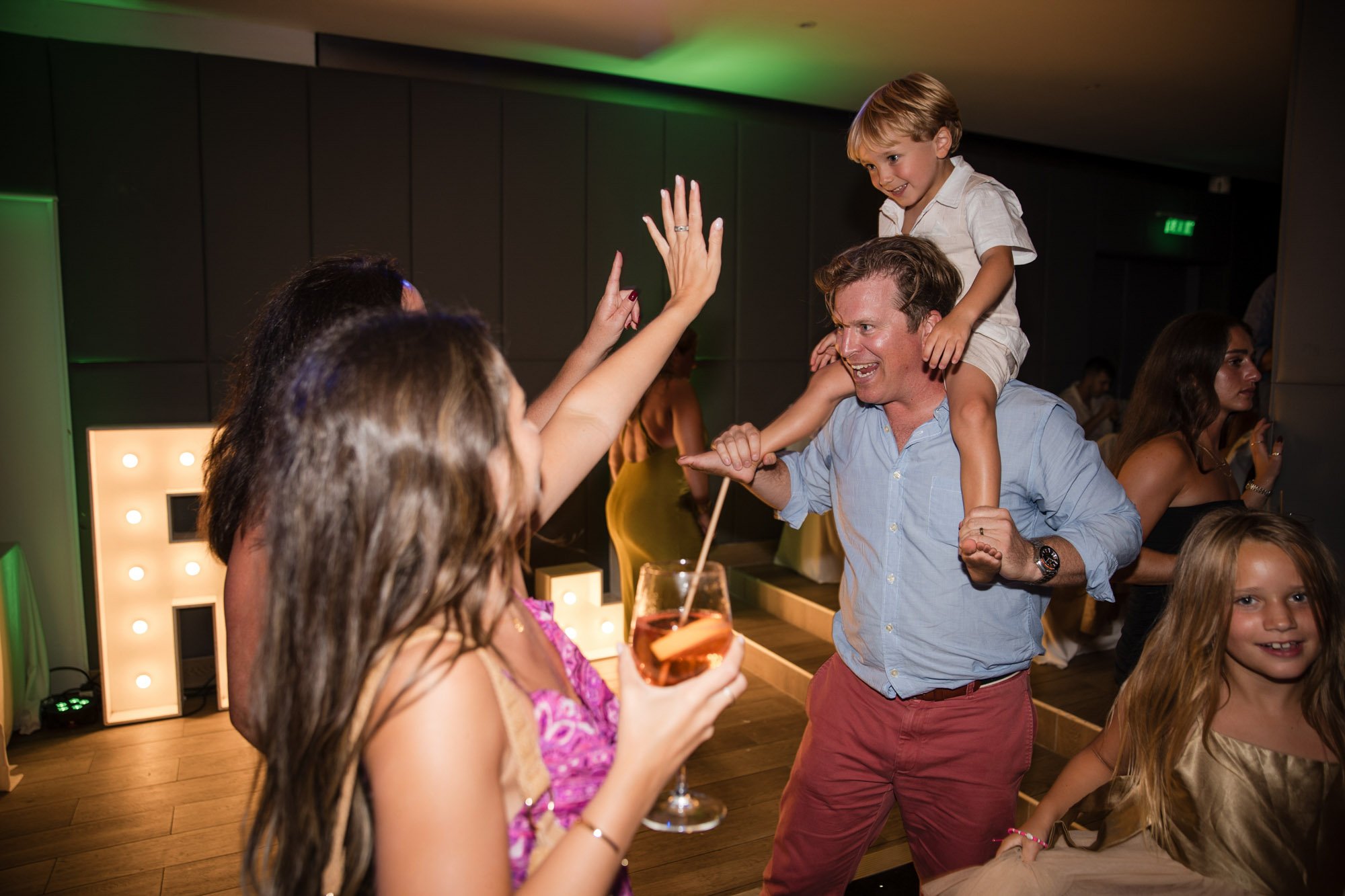 People celebrating at a party; a man with a child on his shoulders dancing and smiling, with women and children around.