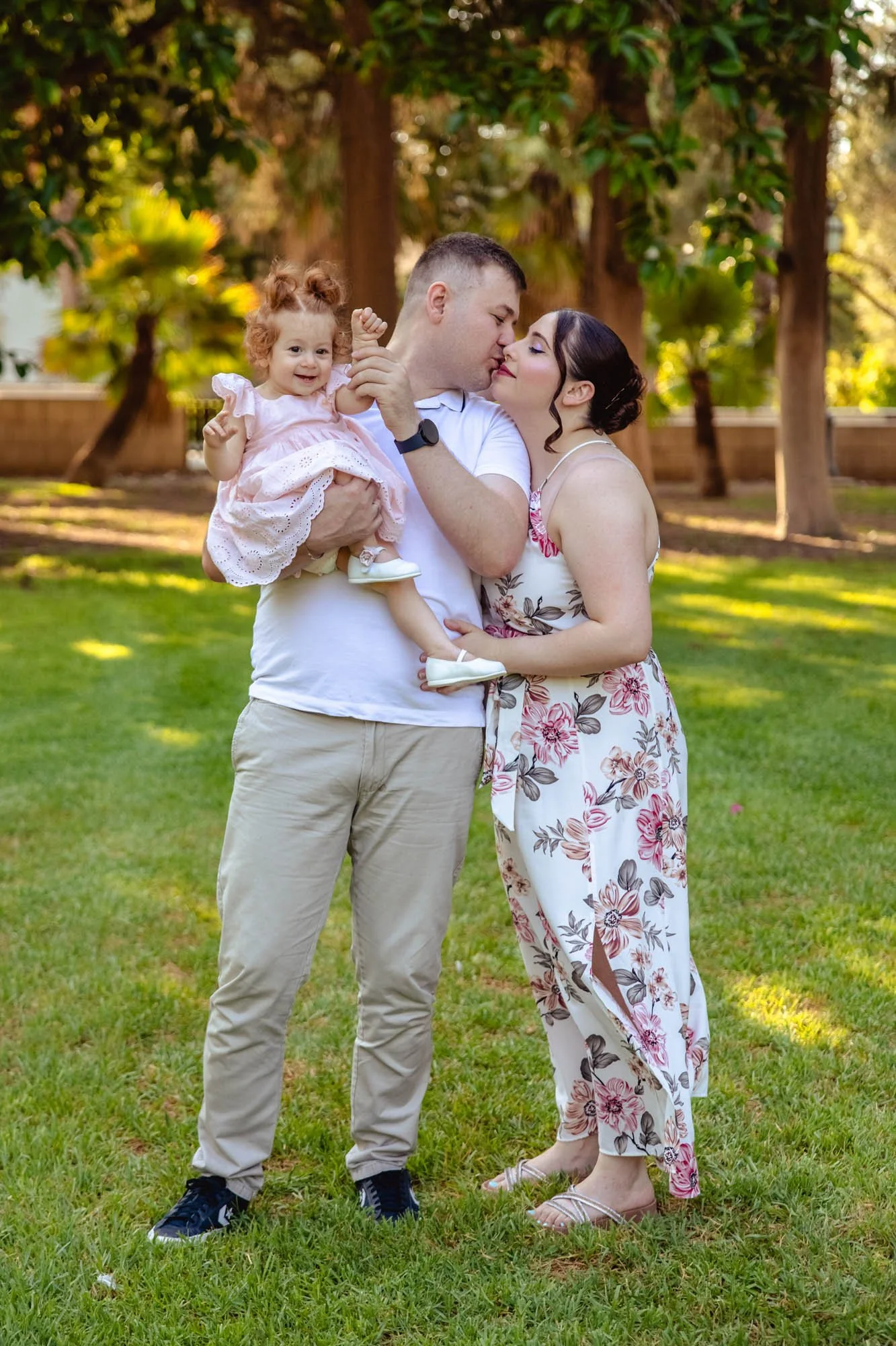 A family of three standing on a grassy lawn surrounded by trees: a man holding a young girl in a pink dress, and a woman in a floral jumpsuit leaning in close to the man. The young girl is smiling and waving.