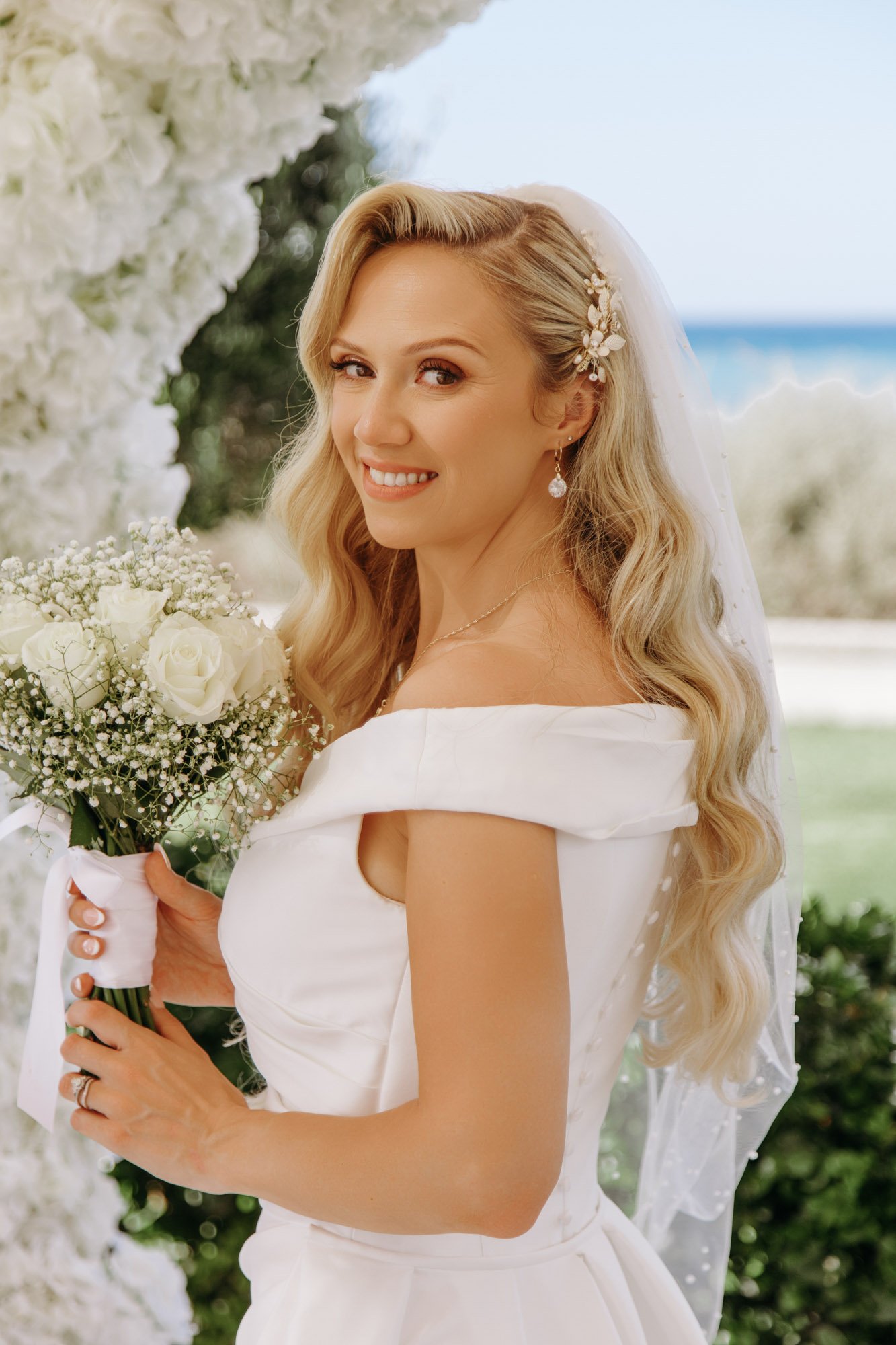 A bride with long blond hair in wedding dress holding a bouquet of white roses and baby's breath, smiling at camera outdoors near the ocean.