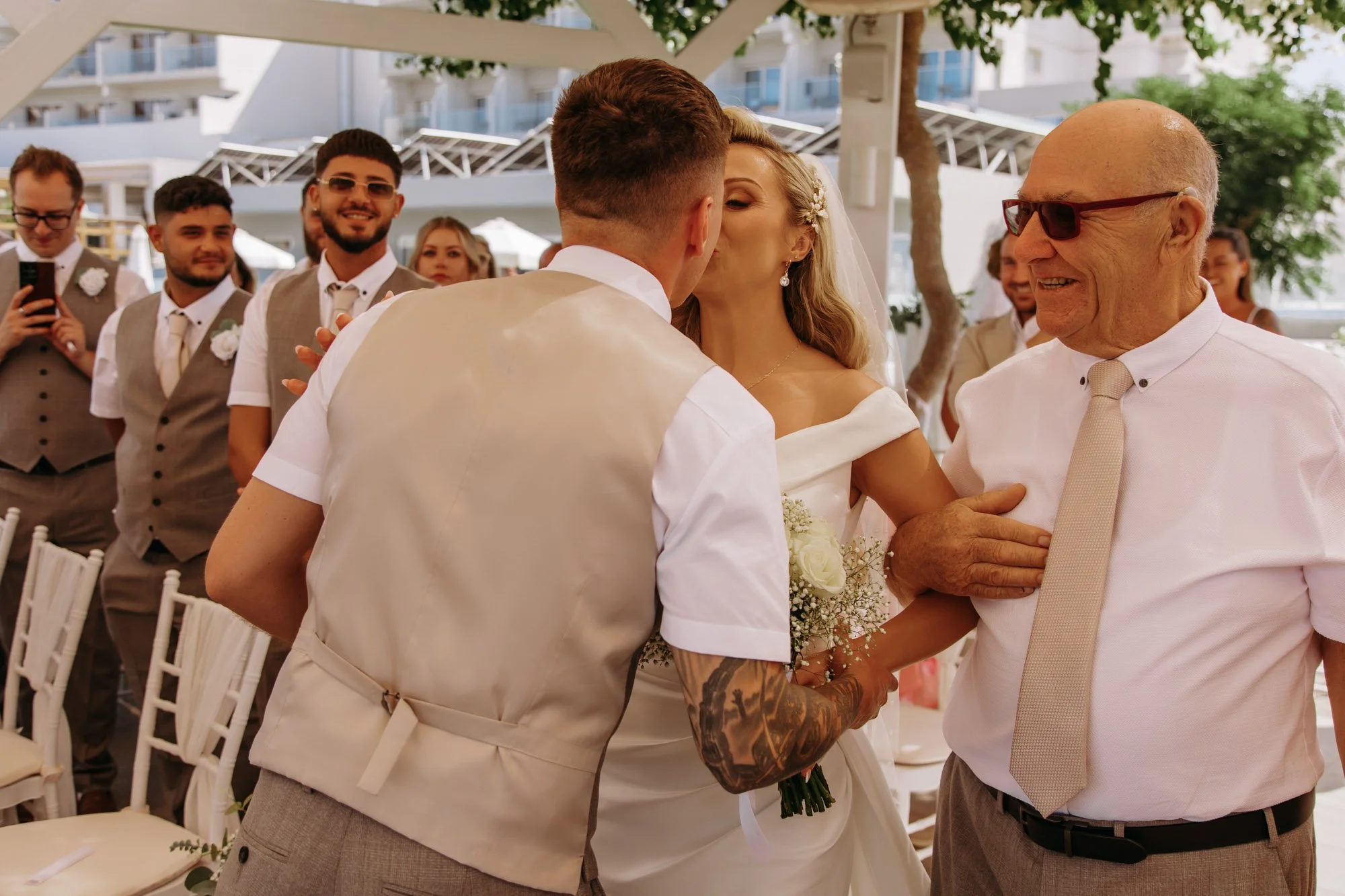 A bride and groom kiss at their wedding ceremony with elderly man smiling nearby, surrounded by groomsmen and guests under a tent.