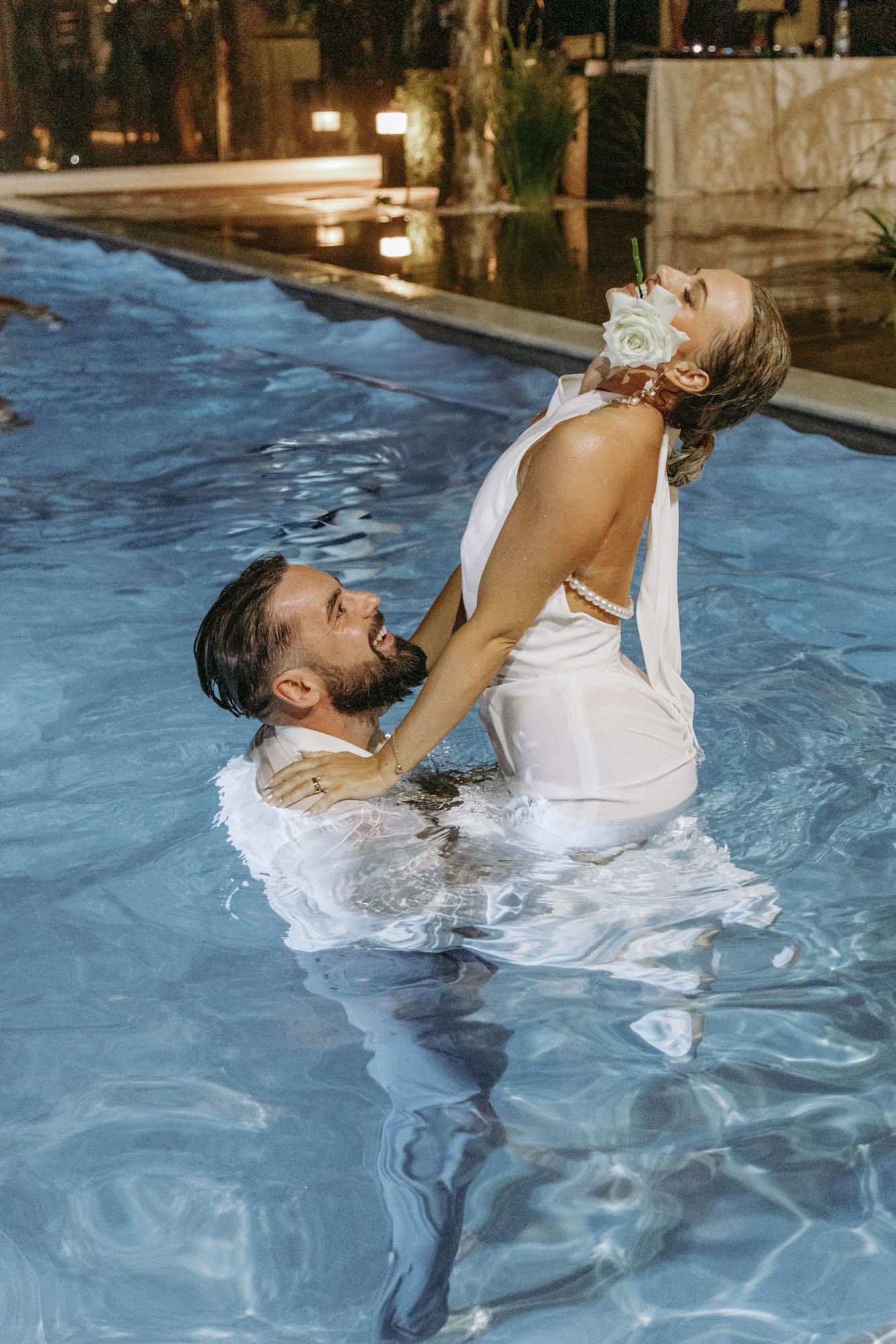 A couple in wedding attire celebrating in a swimming pool at night, with the woman holding a flower in her mouth and the man smiling at her.