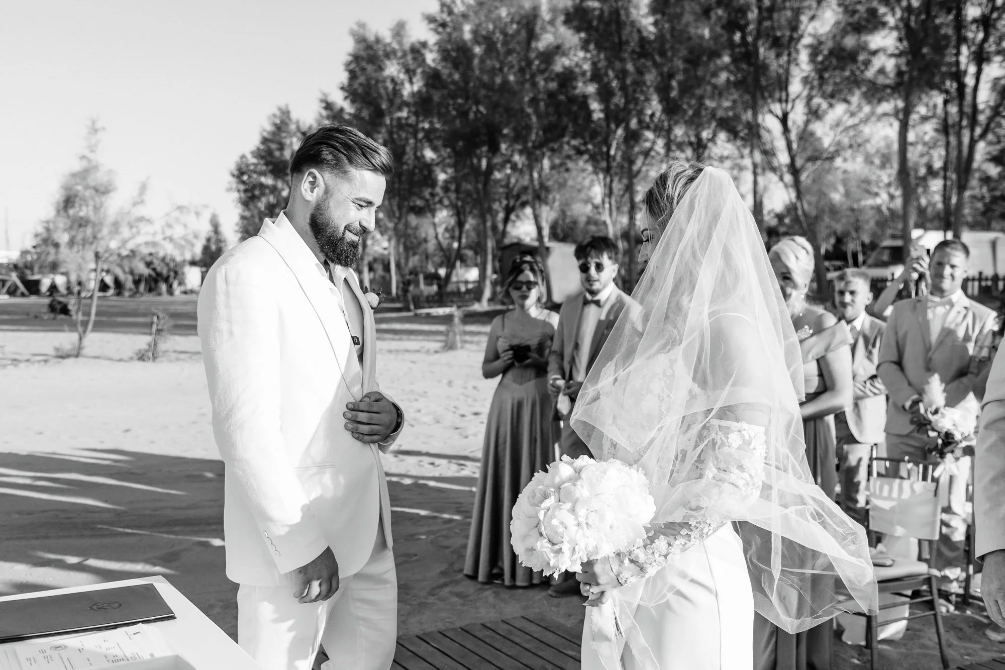 A black and white photo of a wedding ceremony outdoors, showing a groom in a white suit and a bride with a veil holding a bouquet, facing each other with guests watching in the background.