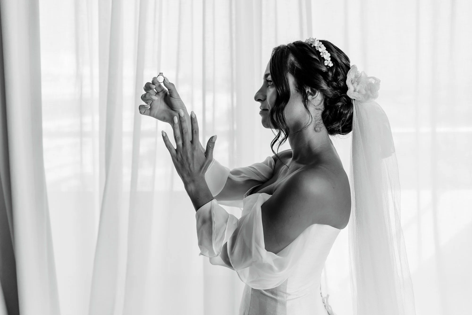 Black and white photo of a bride holding a wedding ring in front of a sheer curtain.