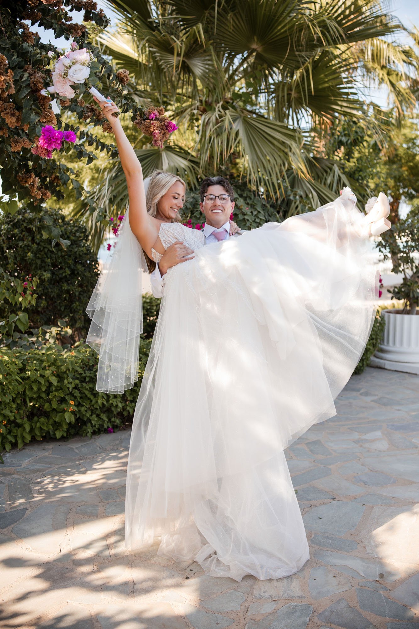 A newlywed couple, with the groom carrying the bride, smiling and laughing outdoors in a garden with palm trees and flowers.