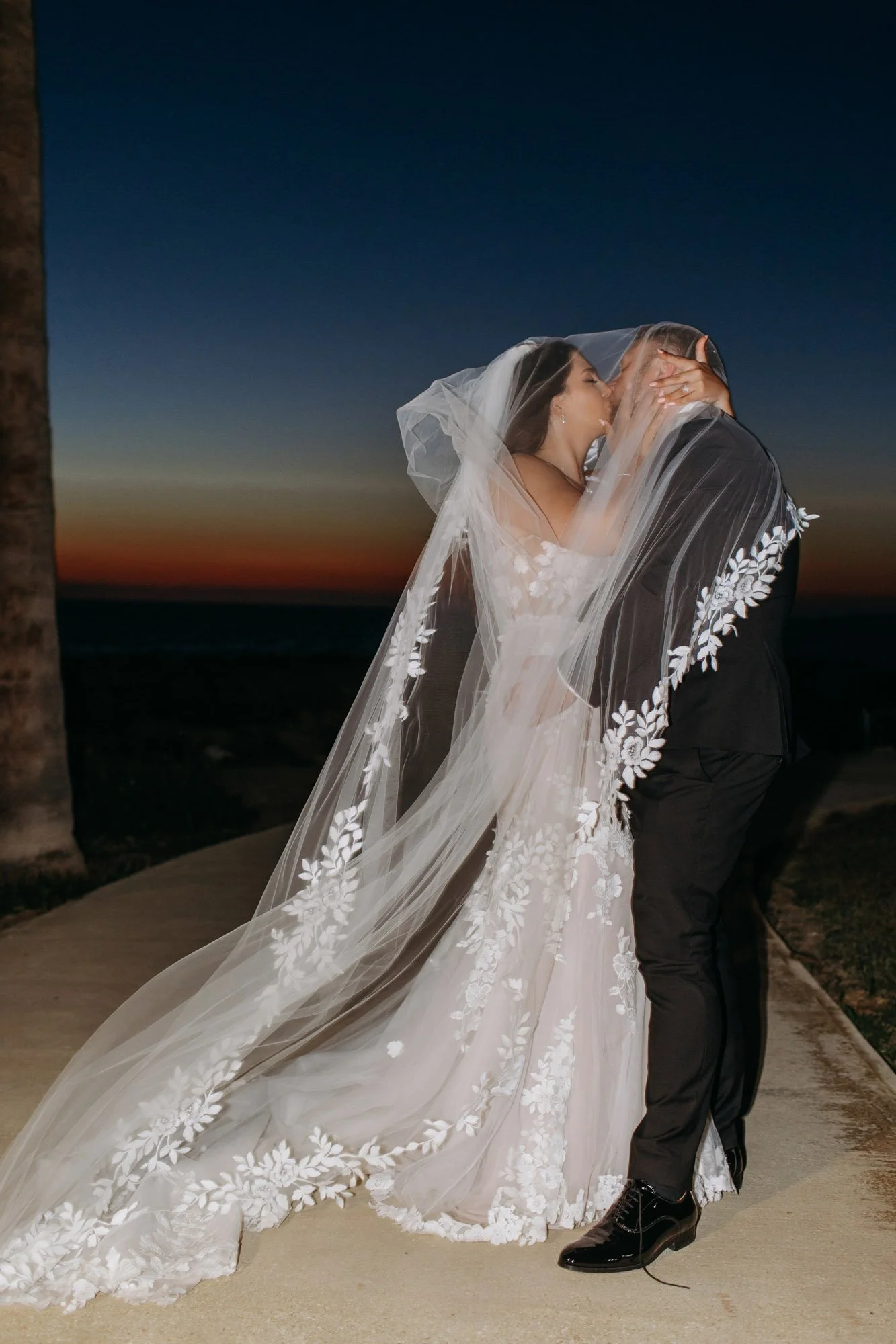A bride and groom sharing a kiss at sunset, with the bride wearing a white wedding gown and veil, and the groom in a black suit, on a pathway near the beach.