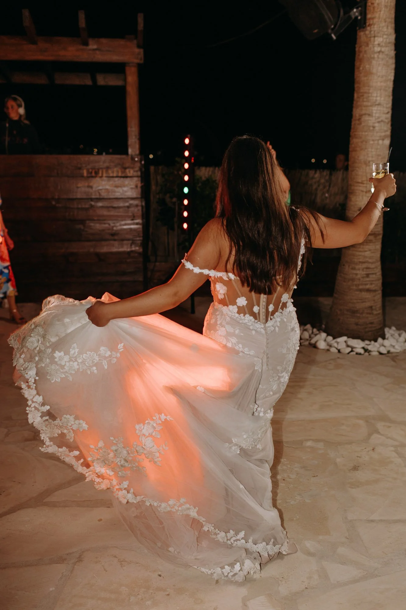 Woman in a white wedding dress dancing at night, holding a glass of champagne, with a DJ in the background and palm trees nearby.
