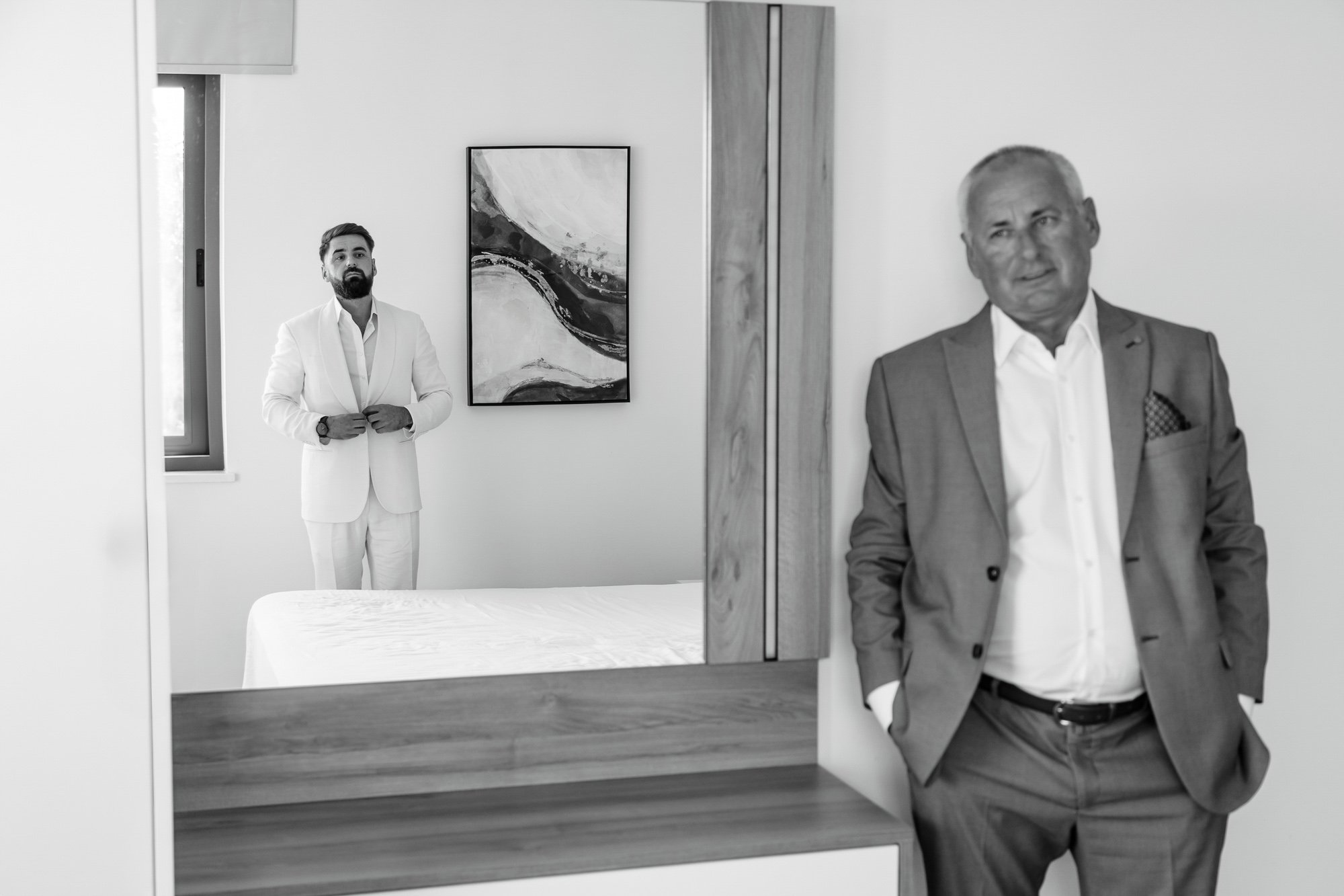 A black-and-white photo of two men in suits in a minimalist, modern room. One man stands in the background near a window, while the other is in the foreground near a mirror, both dressed in formal attire.
