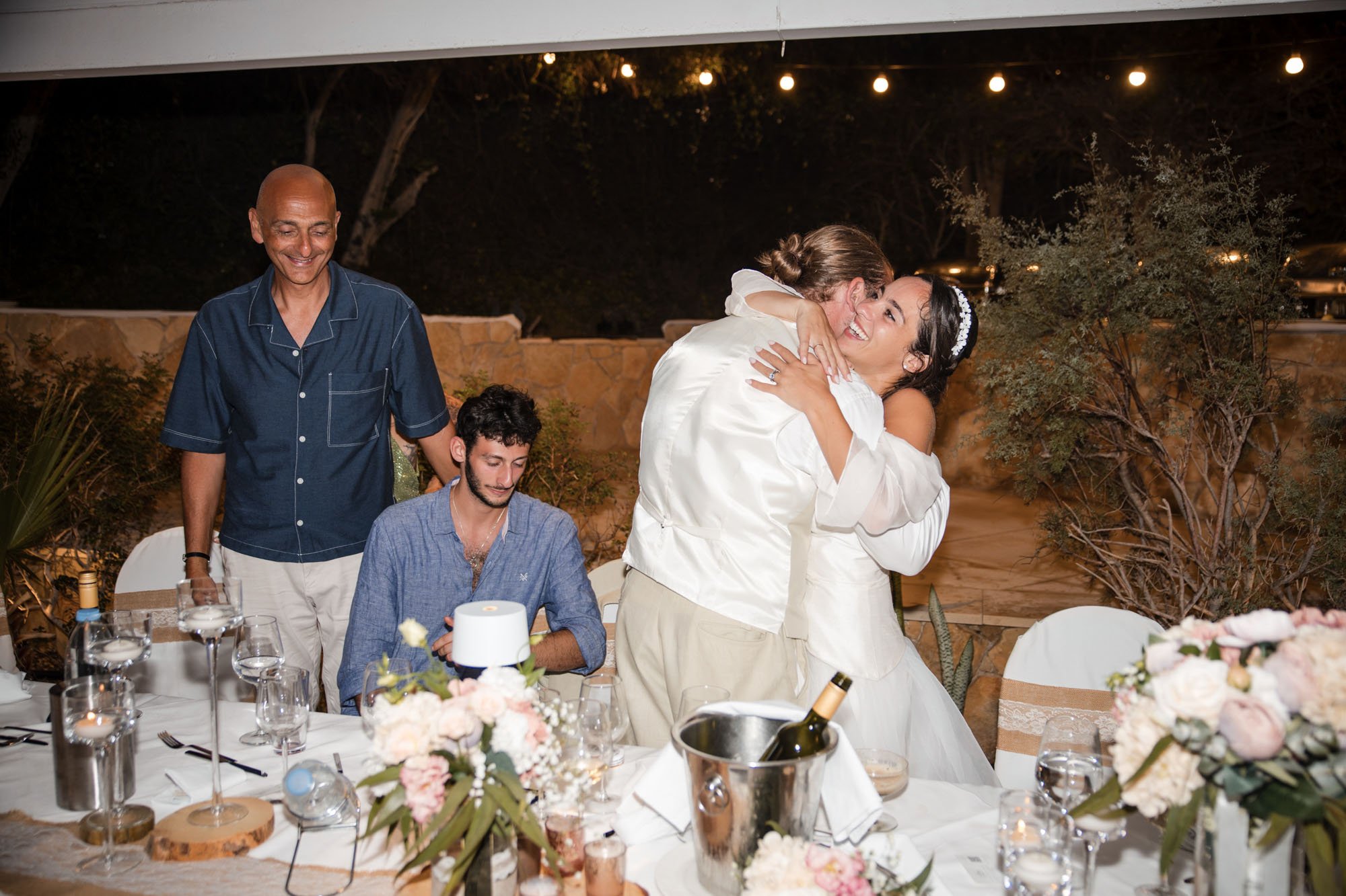 A wedding reception outdoors at night with a table set with flowers, wine, and candles. Two women are hugging and smiling, while two men, one standing and one sitting, observe and smile.
