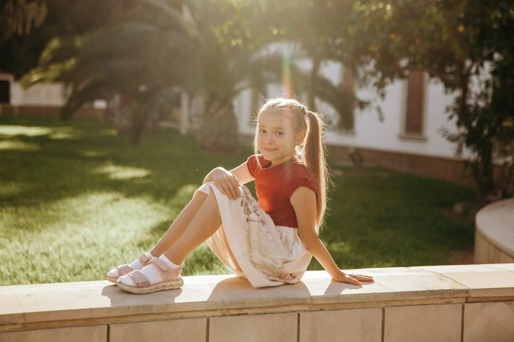 A young girl with long blond hair in pigtails sitting on a low wall outside in the sunlight, smiling, with trees and a white building in the background.