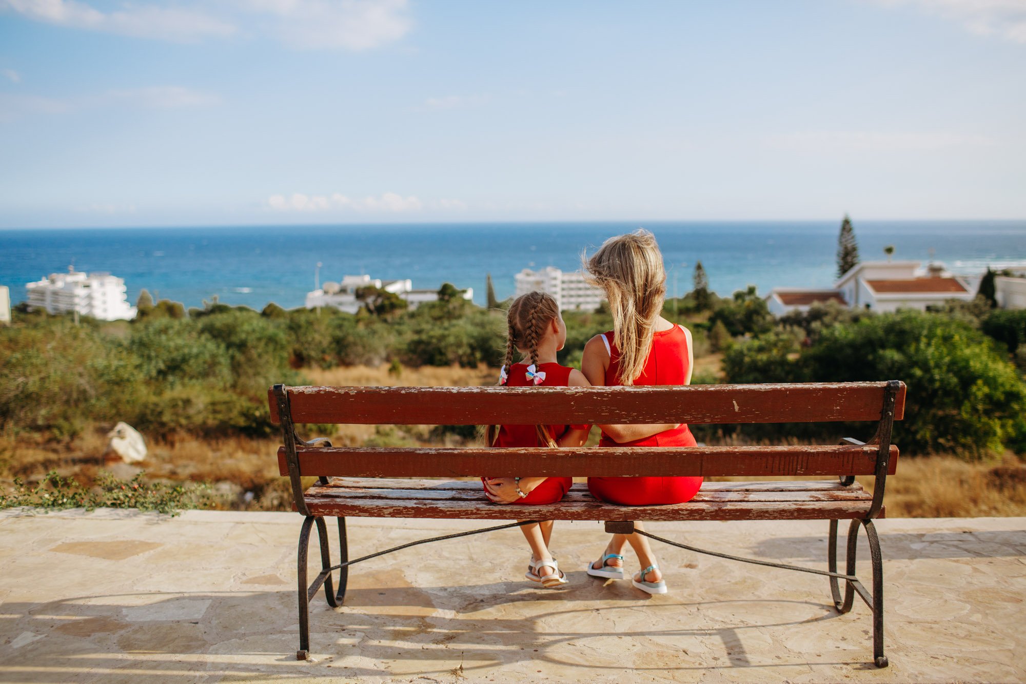 Two girls sitting on a wooden bench facing the ocean, with the background of the sea, blue sky, and houses, enjoying a sunny day.