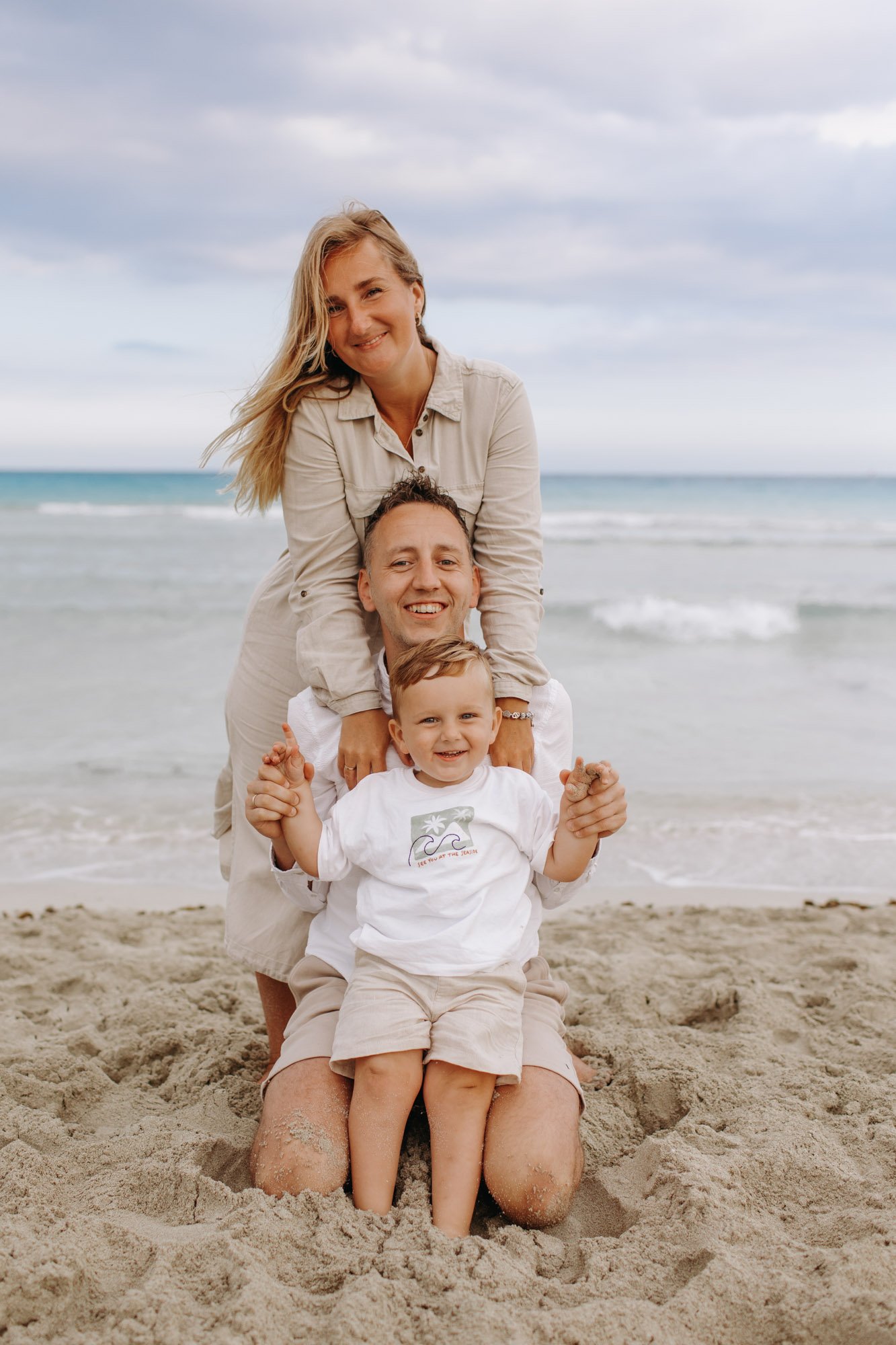 A family of four enjoying a day at the beach, sitting on the sand with the ocean and sky in the background. The mother is smiling, leaning over the father who is kneeling with a child sitting on his lap. All are smiling and wearing casual summer clot