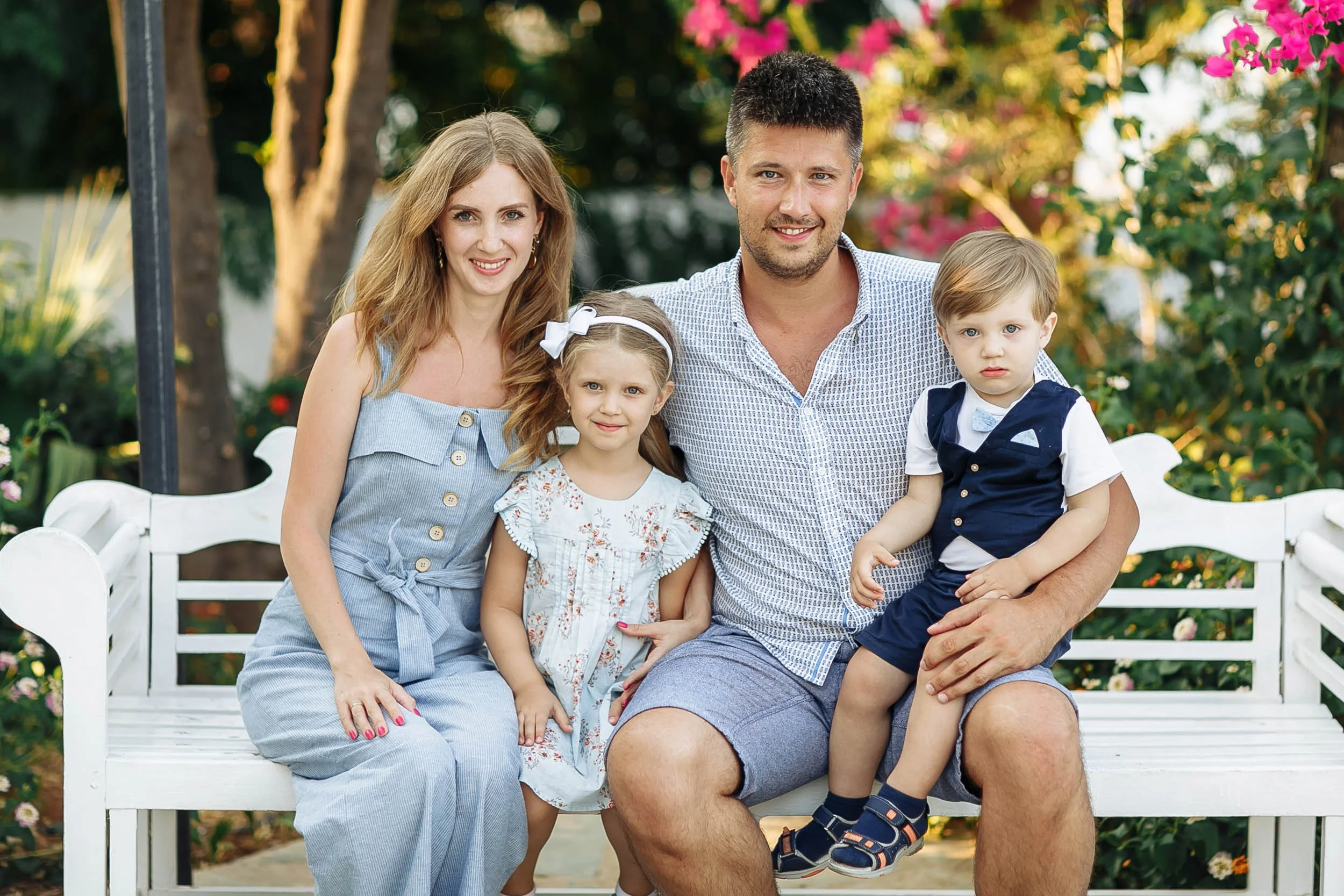 A family of five sitting on a white garden bench outdoors. The family includes a woman with long blonde hair, a man with short dark hair, a young girl with a white bow in her hair, and two young boys. They are surrounded by green foliage and pink flo