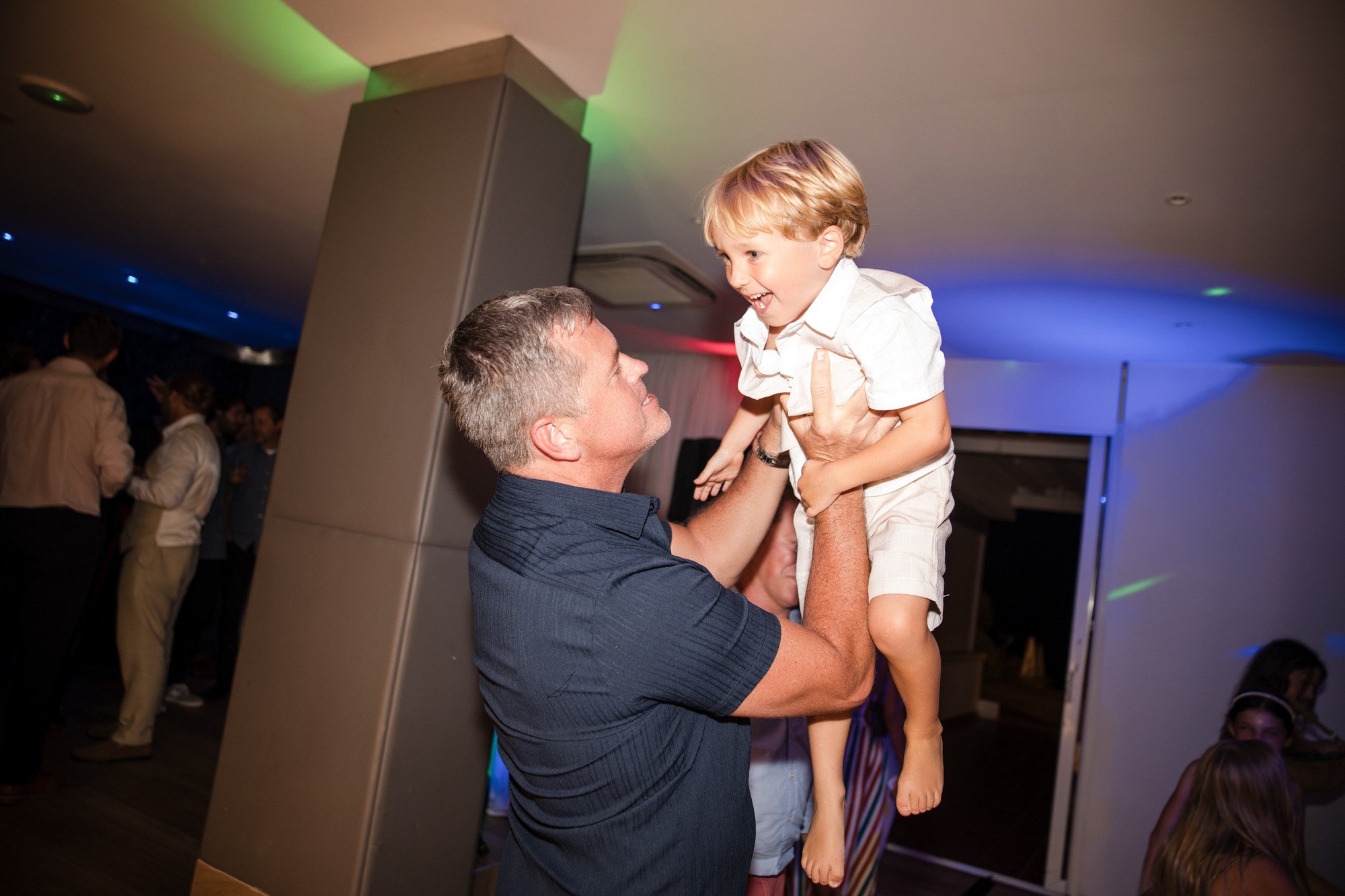 A man lifting a young boy in the air during a celebration or party, with colorful lights and other people in the background.