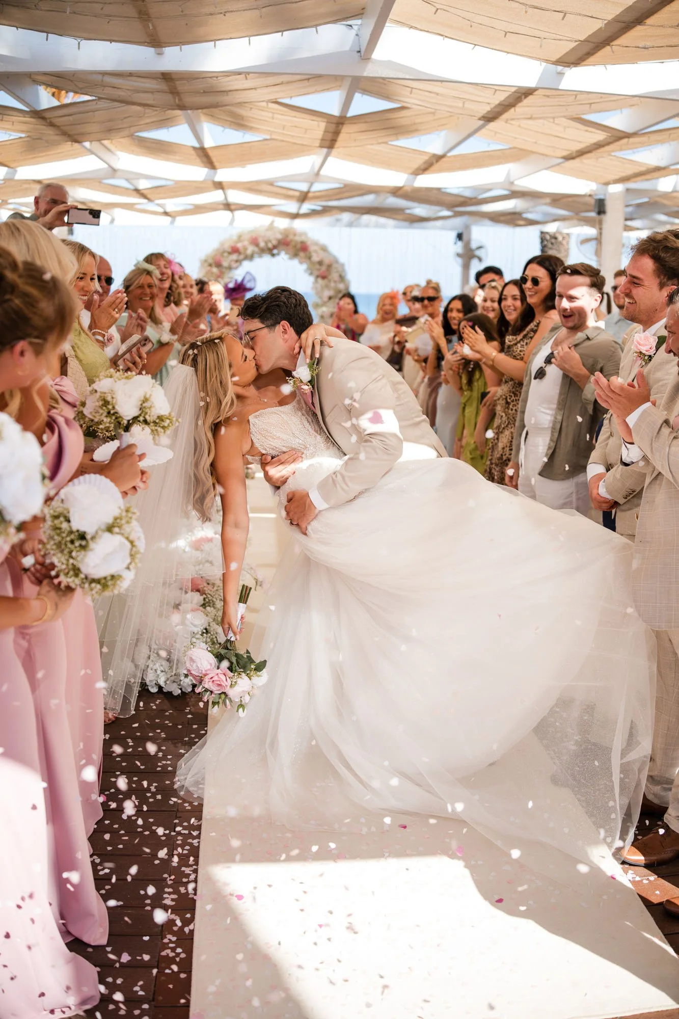 A newlywed couple sharing a kiss as they dip during their wedding ceremony, surrounded by cheering guests under a decorated canopy.
