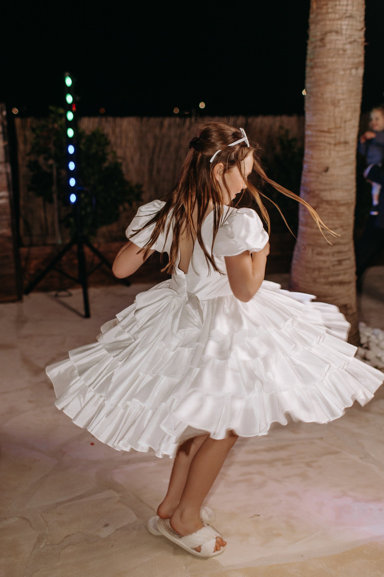 A young girl in a white dress spinning on a dance floor at night, with a palm tree and string lights in the background.