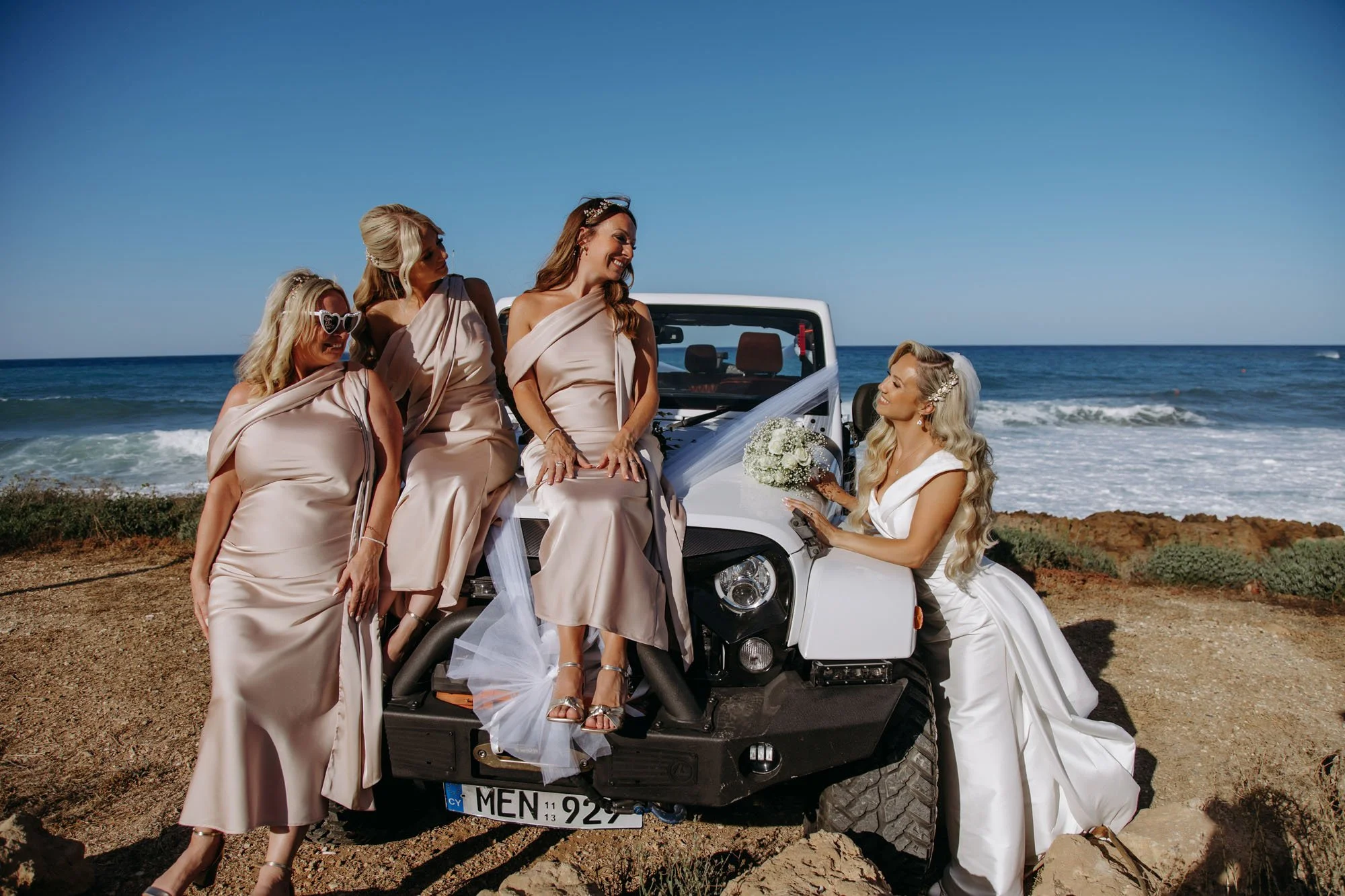 Bride in white dress with long blonde hair and veil holding bouquet, surrounded by four women in beige dresses on a white off-road vehicle near the ocean.
