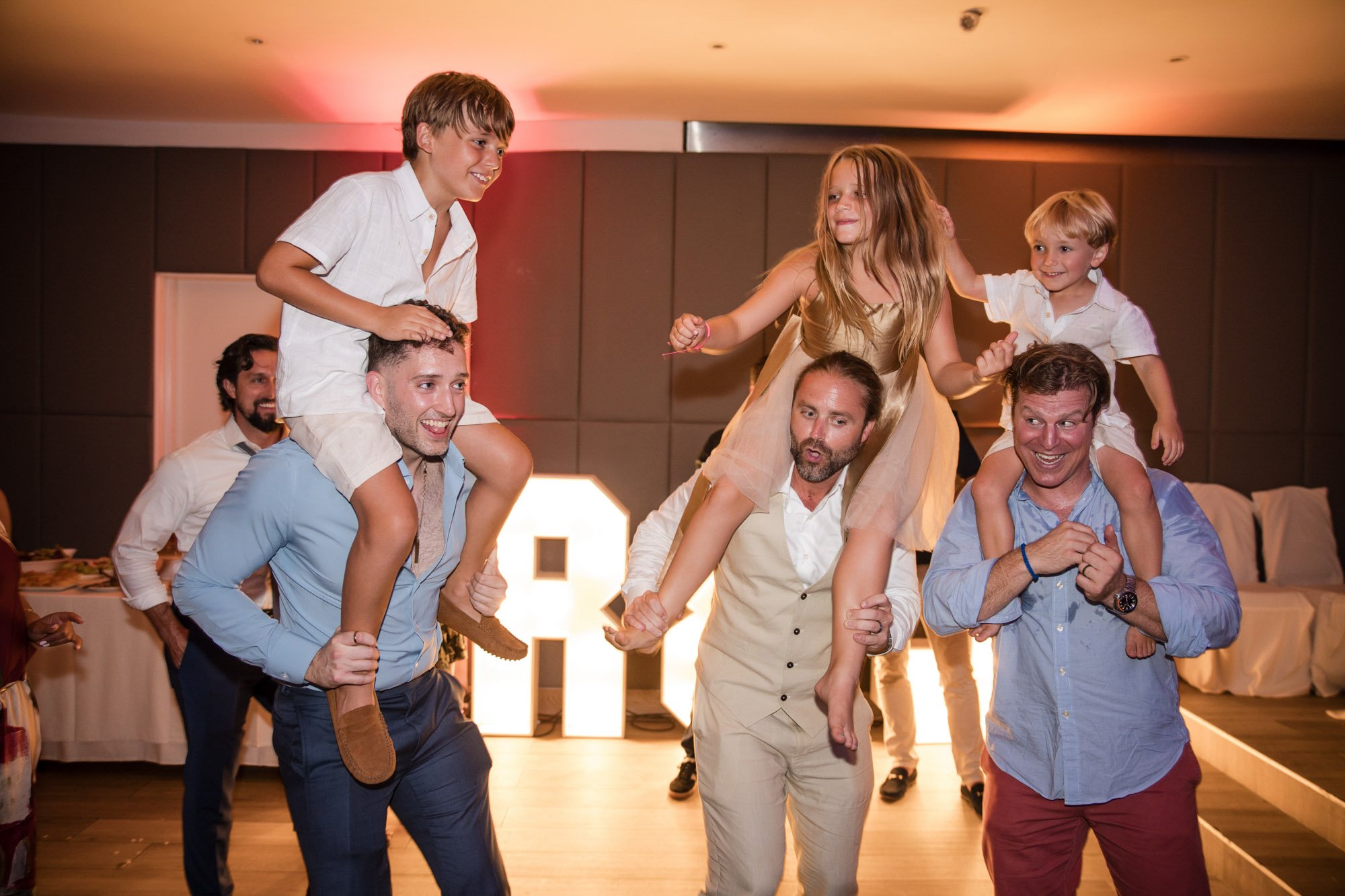 Group of adults and children joyfully celebrating and dancing at a party, with some children riding on the shoulders of adults.