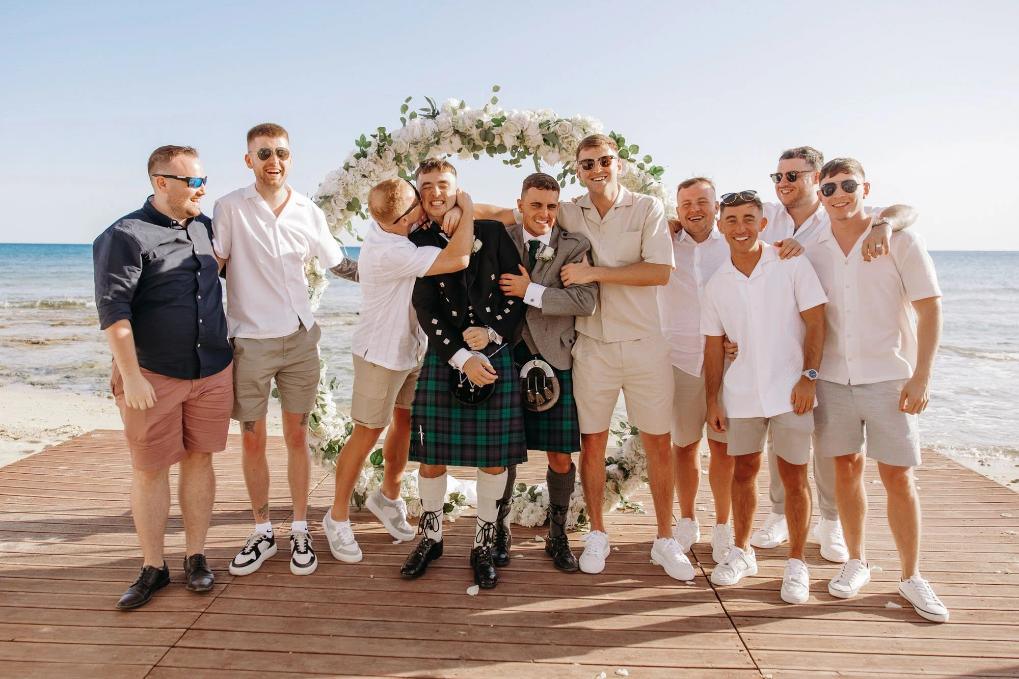 Group of men at a beach wedding, standing on a wooden platform with the ocean in the background, celebrating in casual attire with sunglasses and smiling.