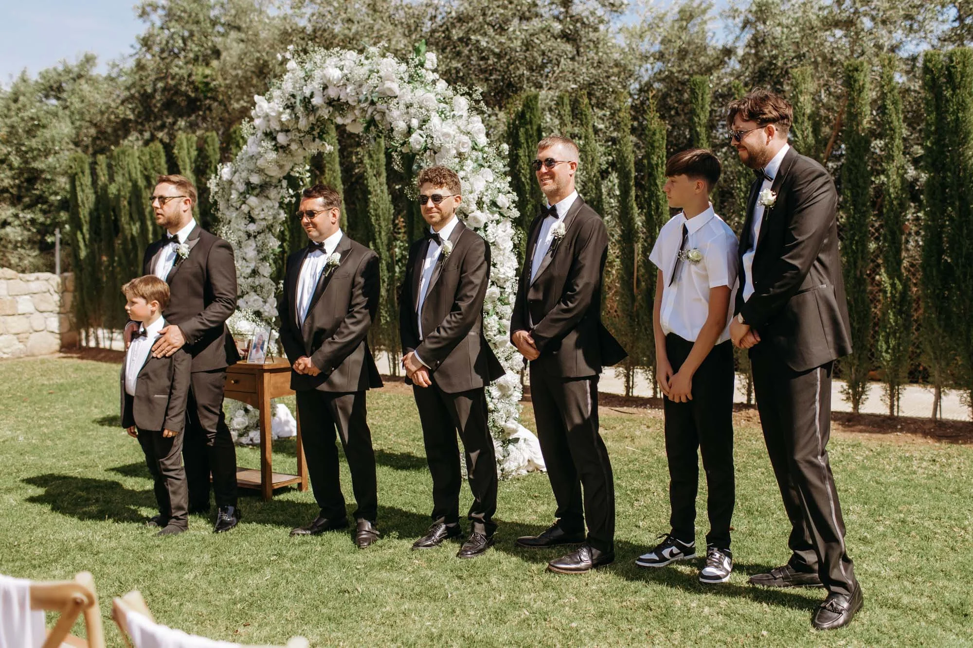 Group of groom and groomsmen standing outdoors at wedding ceremony in front of floral arch with greenery background.